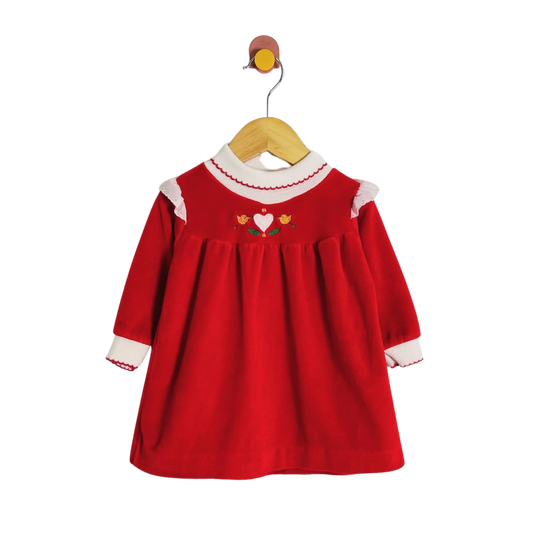 A red long-sleeve baby dress with white ruffled trim at the collar and cuffs, featuring a small embroidered heart and flowers on the chest, hanging on a wooden hanger against a white background.