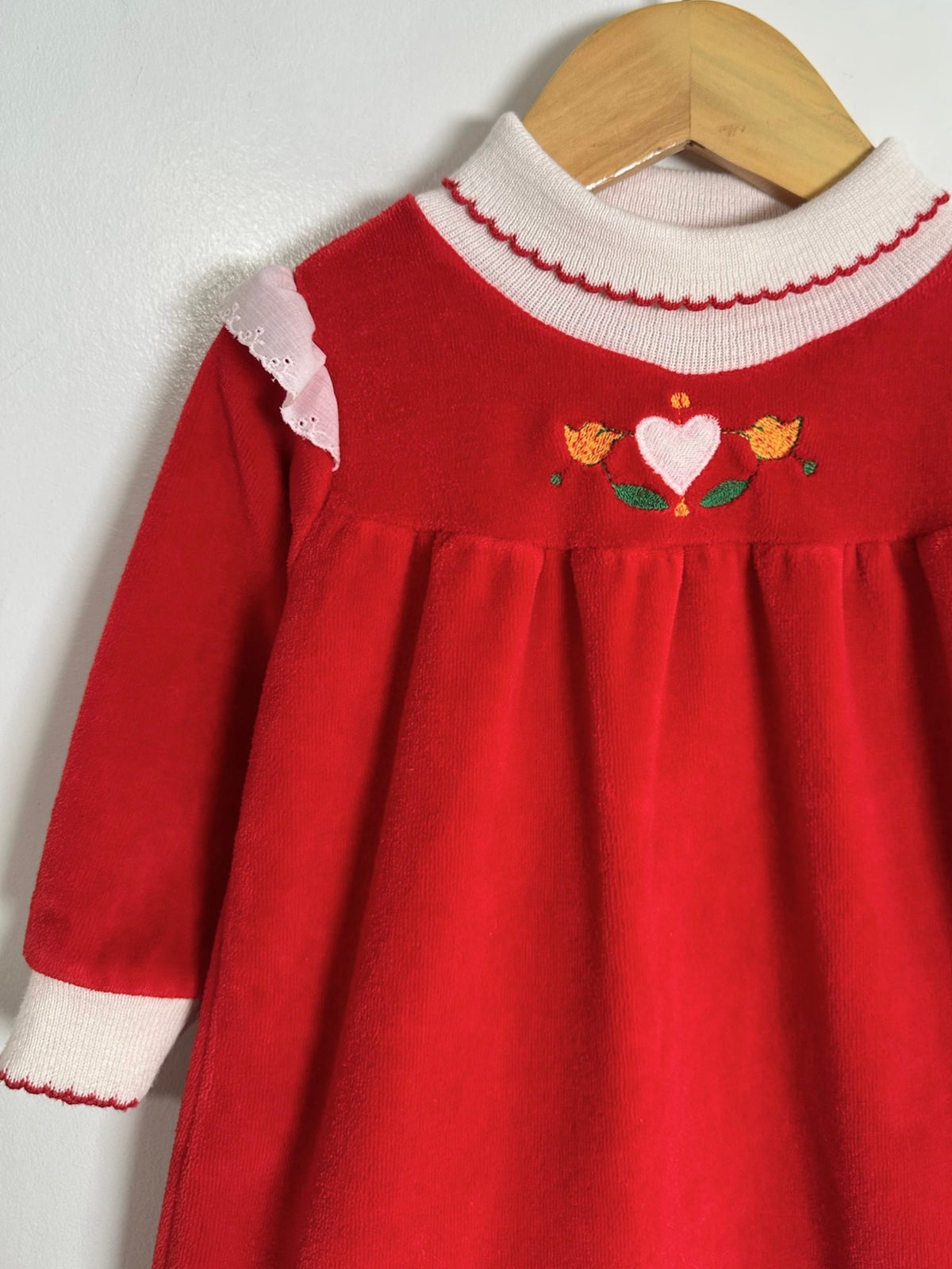 A red long-sleeve dress with a white ribbed mock neck and cuffs, featuring a small embroidered heart and flowers on the chest, hanging on a wooden hanger against a white wall.