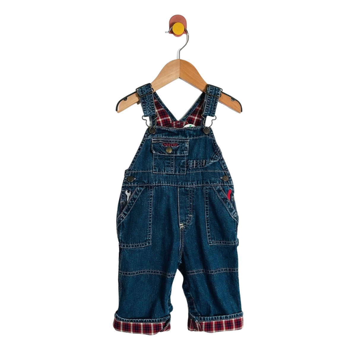 A pair of baby denim overall  with red and black plaid lining under the straps and at the cuffed hems, hanging on a wooden hanger against a white background.