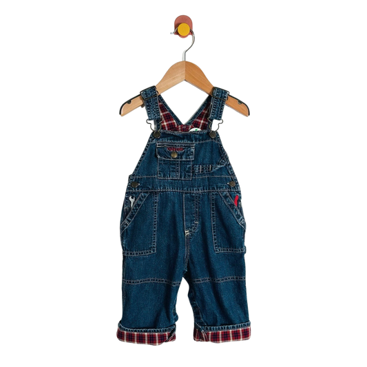 A pair of baby denim overall  with red and black plaid lining under the straps and at the cuffed hems, hanging on a wooden hanger against a white background.