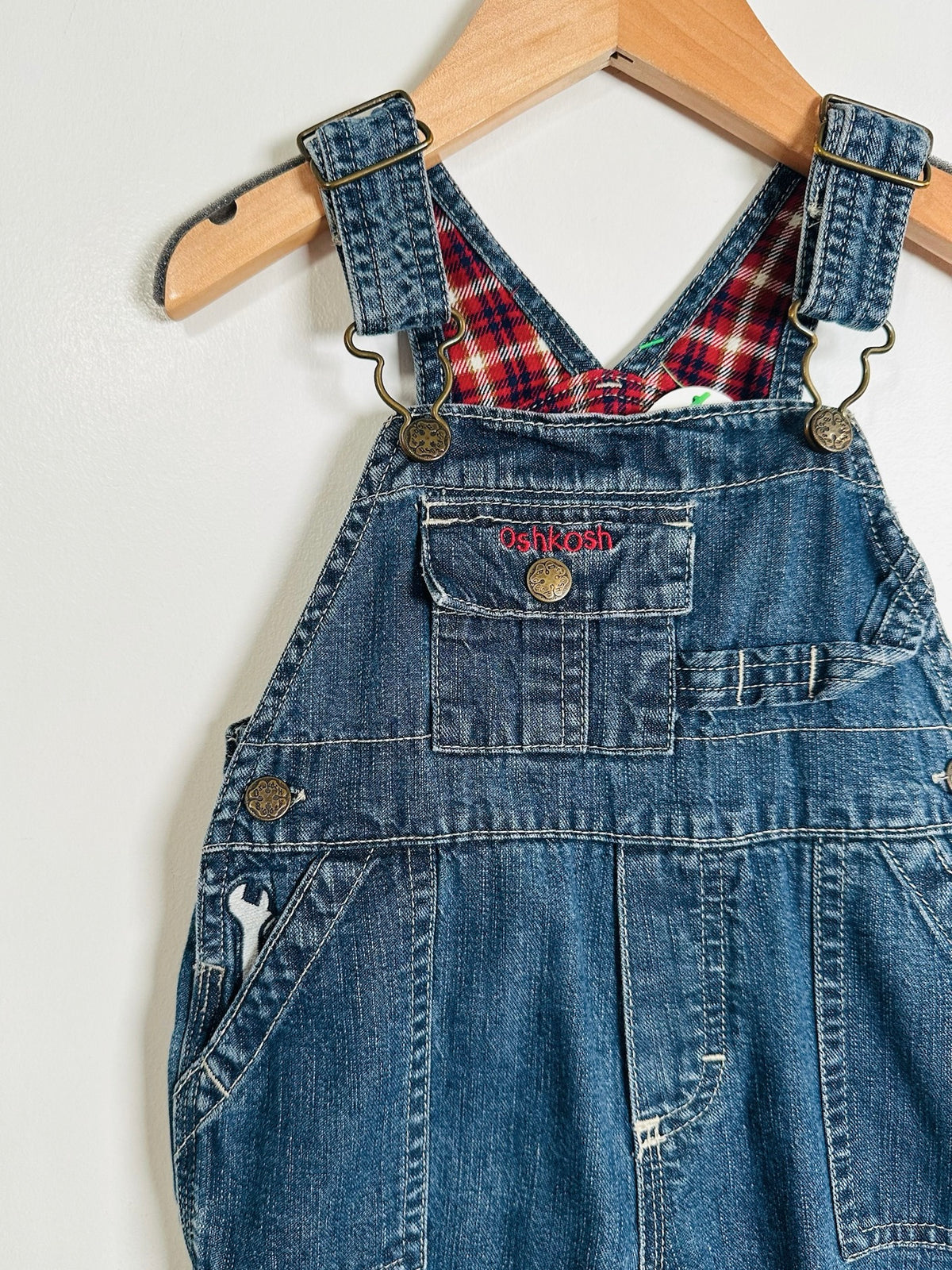 A pair of blue denim baby overalls hanging on a wooden hanger against a plain white wall. The overalls feature adjustable straps with brass-tone metal hardware, a front chest pocket with a snap closure, and a red and black plaid lining visible at the neckline.