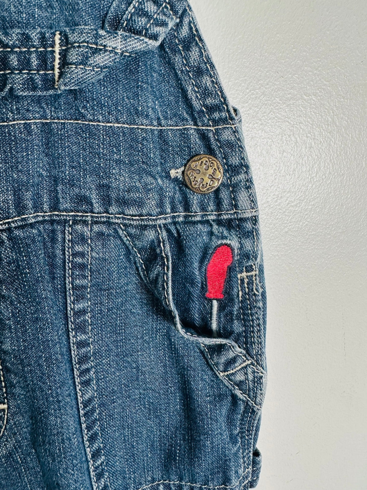 Close-up of a pair of blue denim overalls showing the pocket detail with a red embroidered patch and a brass button.