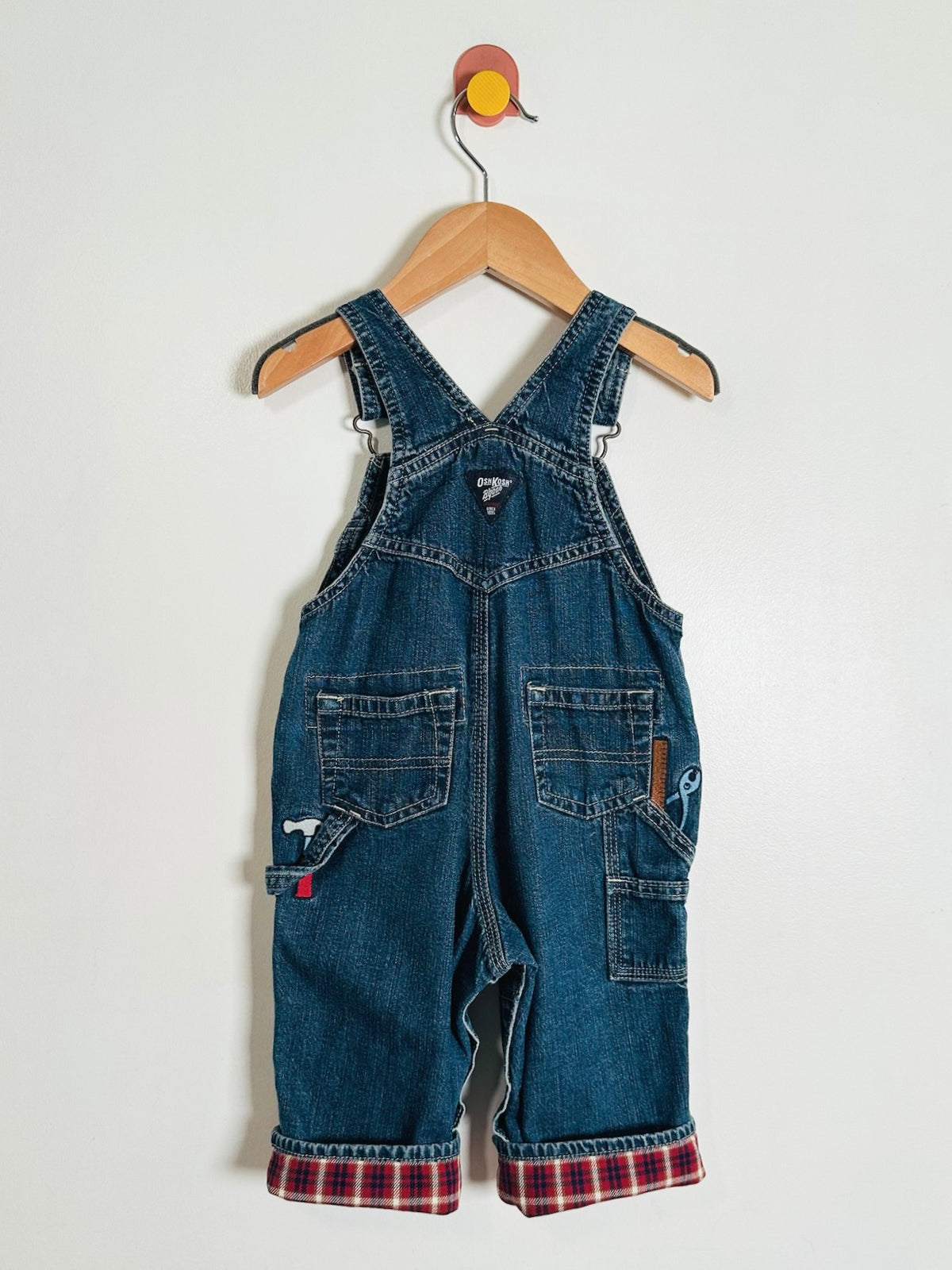 A pair of toddler denim bib overalls hanging on a wooden hanger against a plain white wall. The overalls are made of medium blue denim with adjustable shoulder straps, front and back patch pockets, and a red and blue plaid lining visible at the cuffed hems.