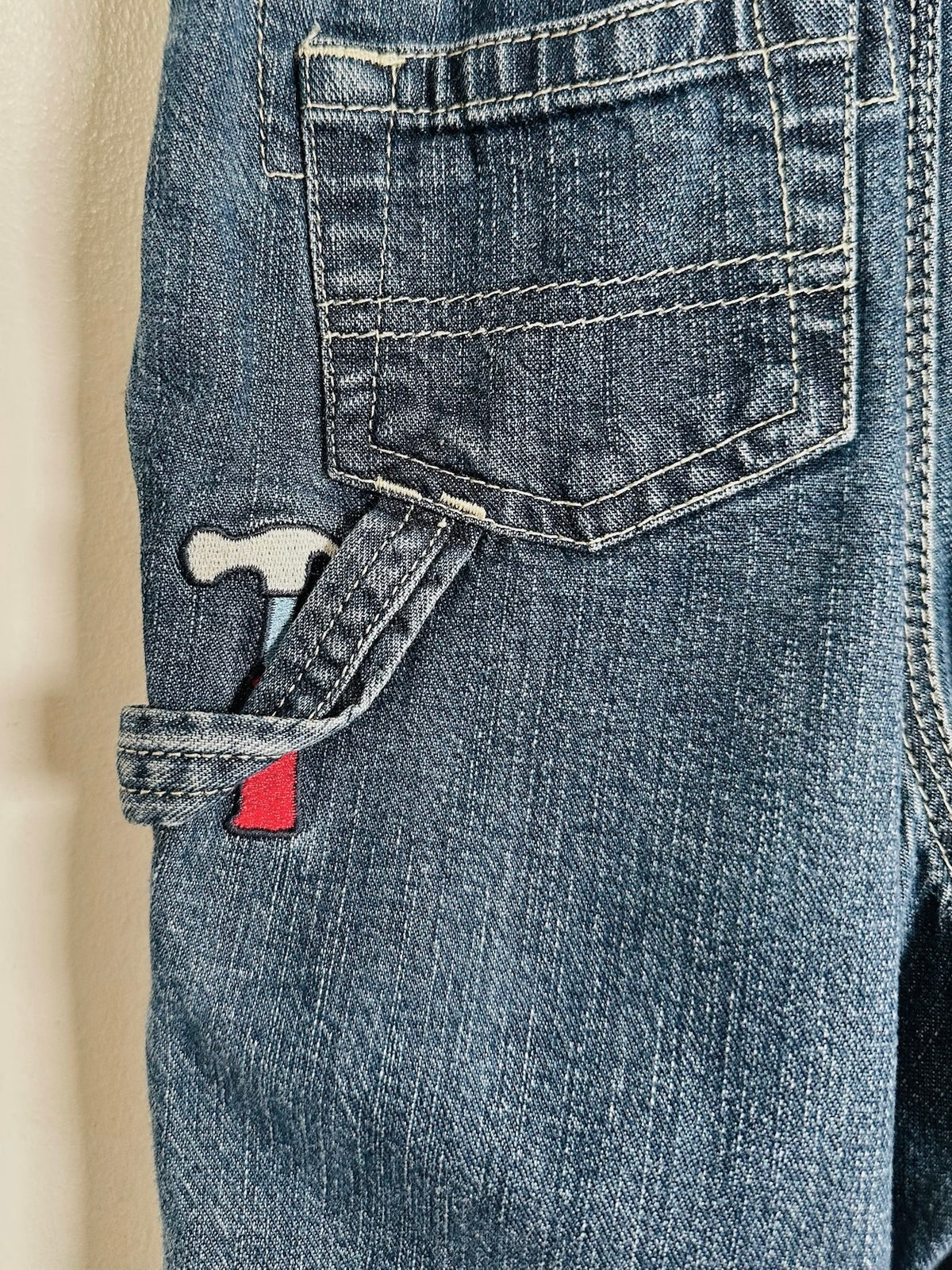 Close-up of a pair of blue denim jeans showing the back pocket, cargo pocket with a hammer patch, and a small red fabric tag.