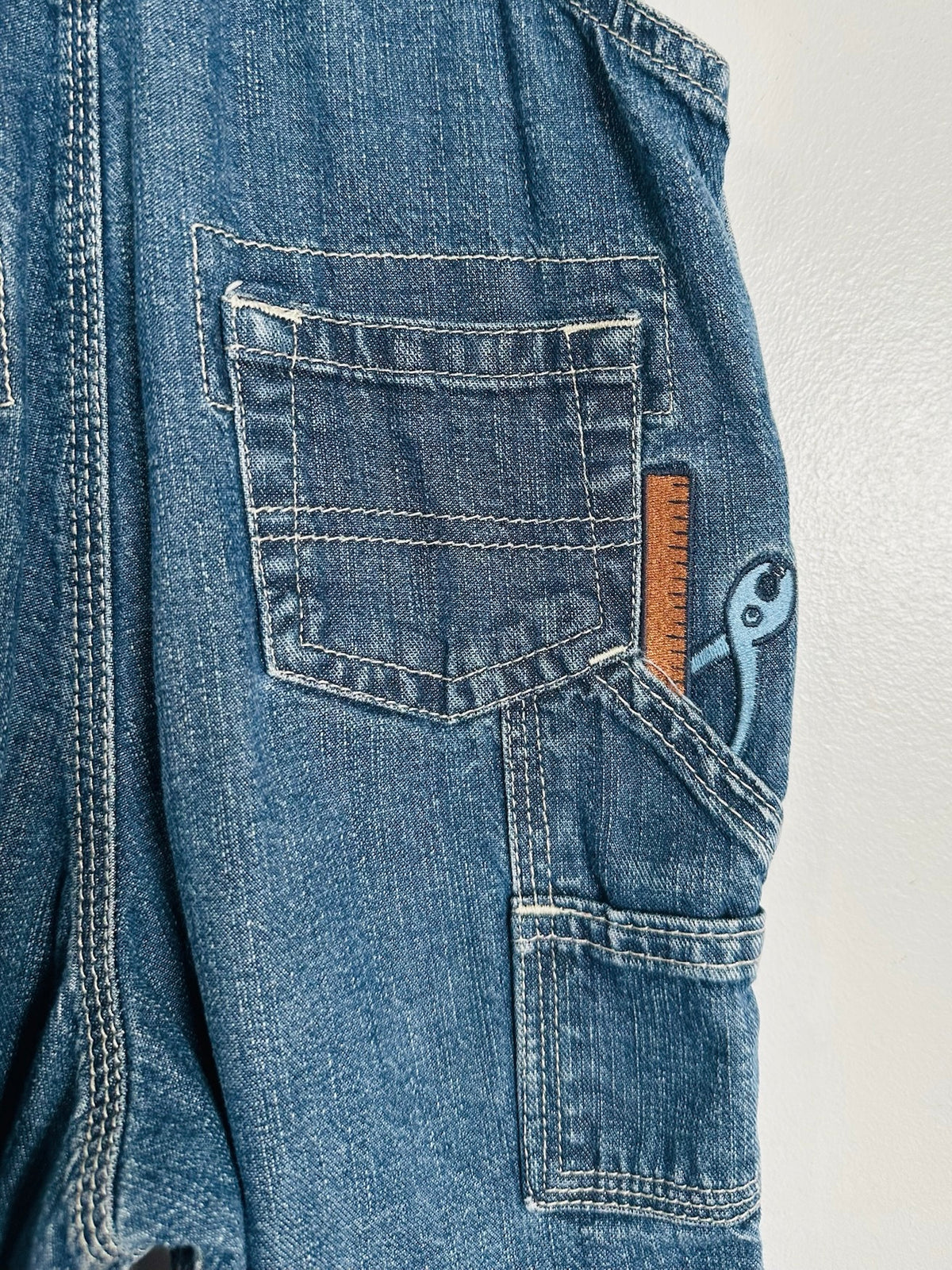 Close-up of a blue denim work jacket or bib with front and side cargo pockets, showing a small blue wrench patch and a brown leather measurement patch on the pocket.