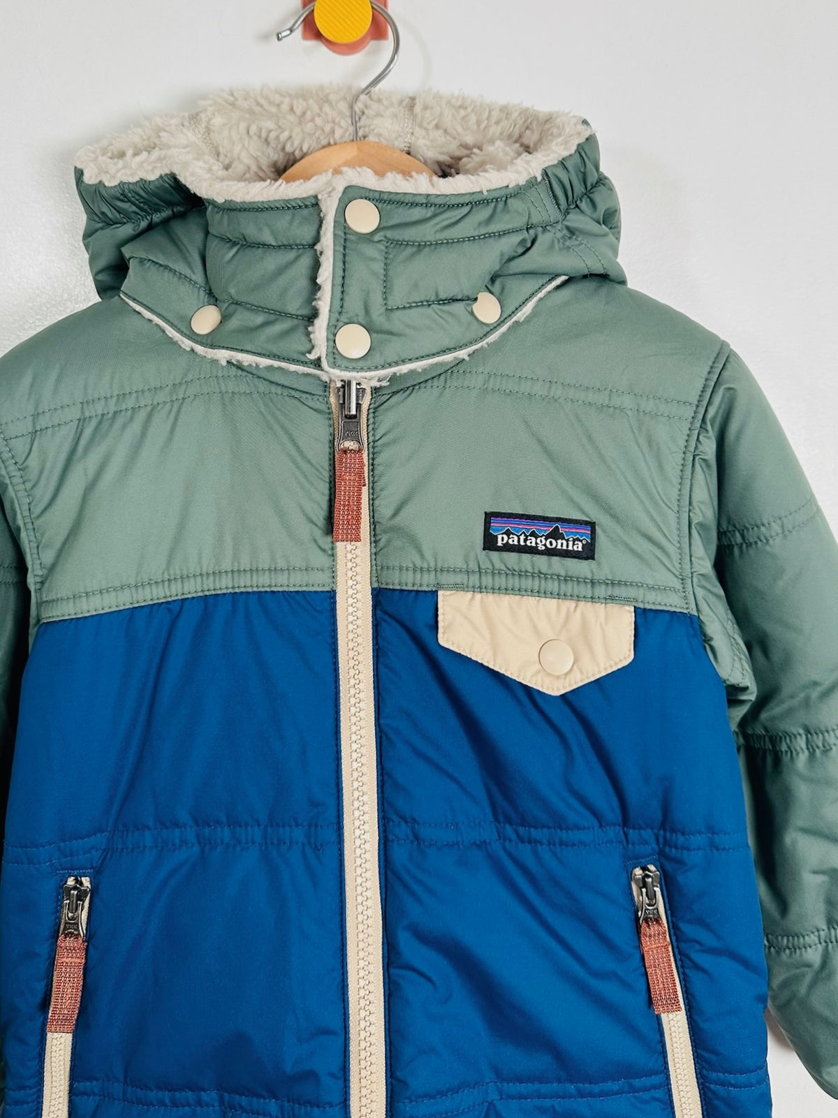 A Patagonia hooded puffer jacket hanging on a wooden hanger against a plain white wall. The jacket features a sage green upper section, a royal blue lower section, a beige faux-fur lined hood, and a beige chest pocket.