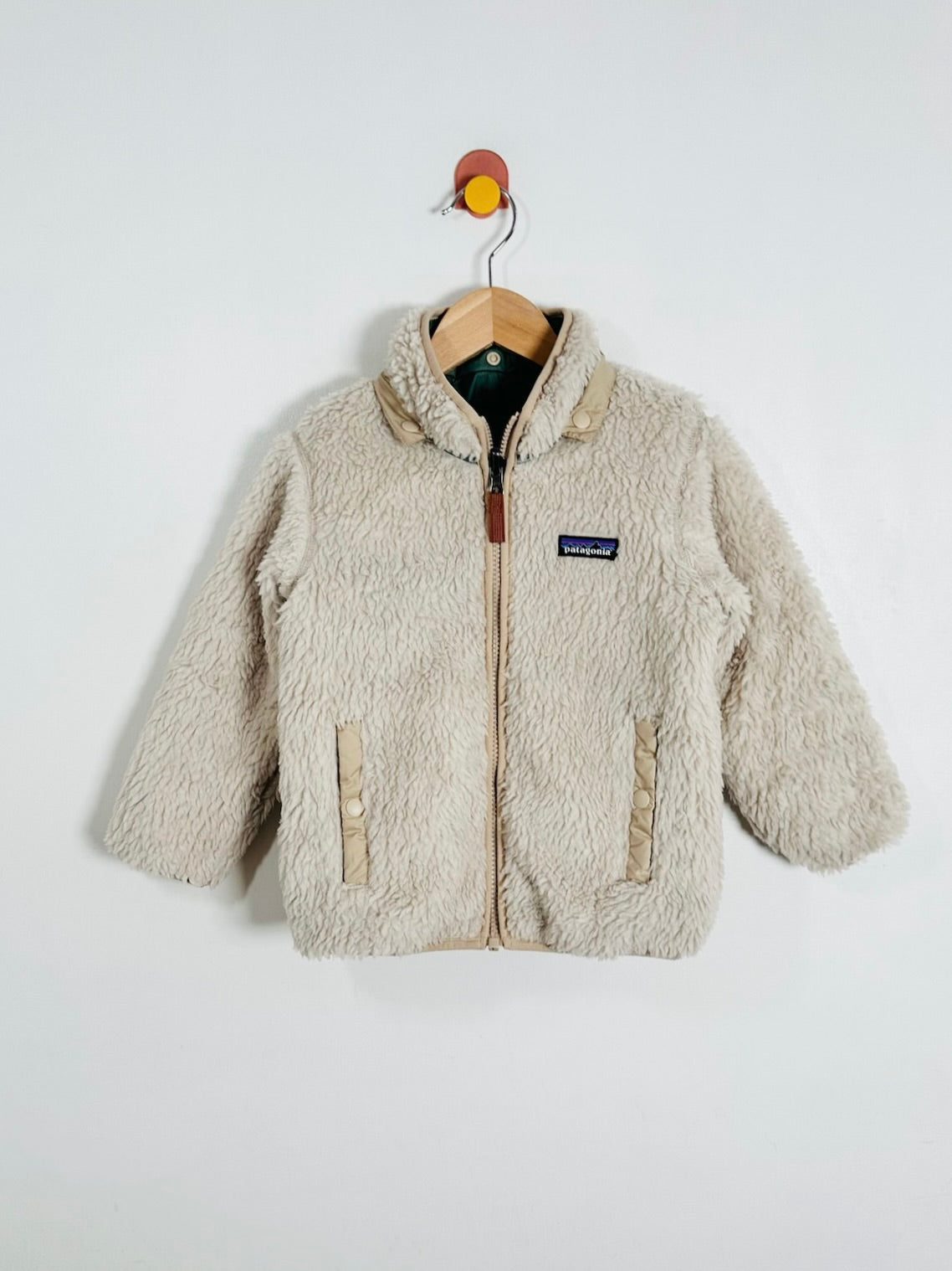 A beige sherpa fleece zip-up jacket with a stand collar, hanging on a wooden hanger against a plain white wall. A small Patagonia logo patch is visible on the left chest.