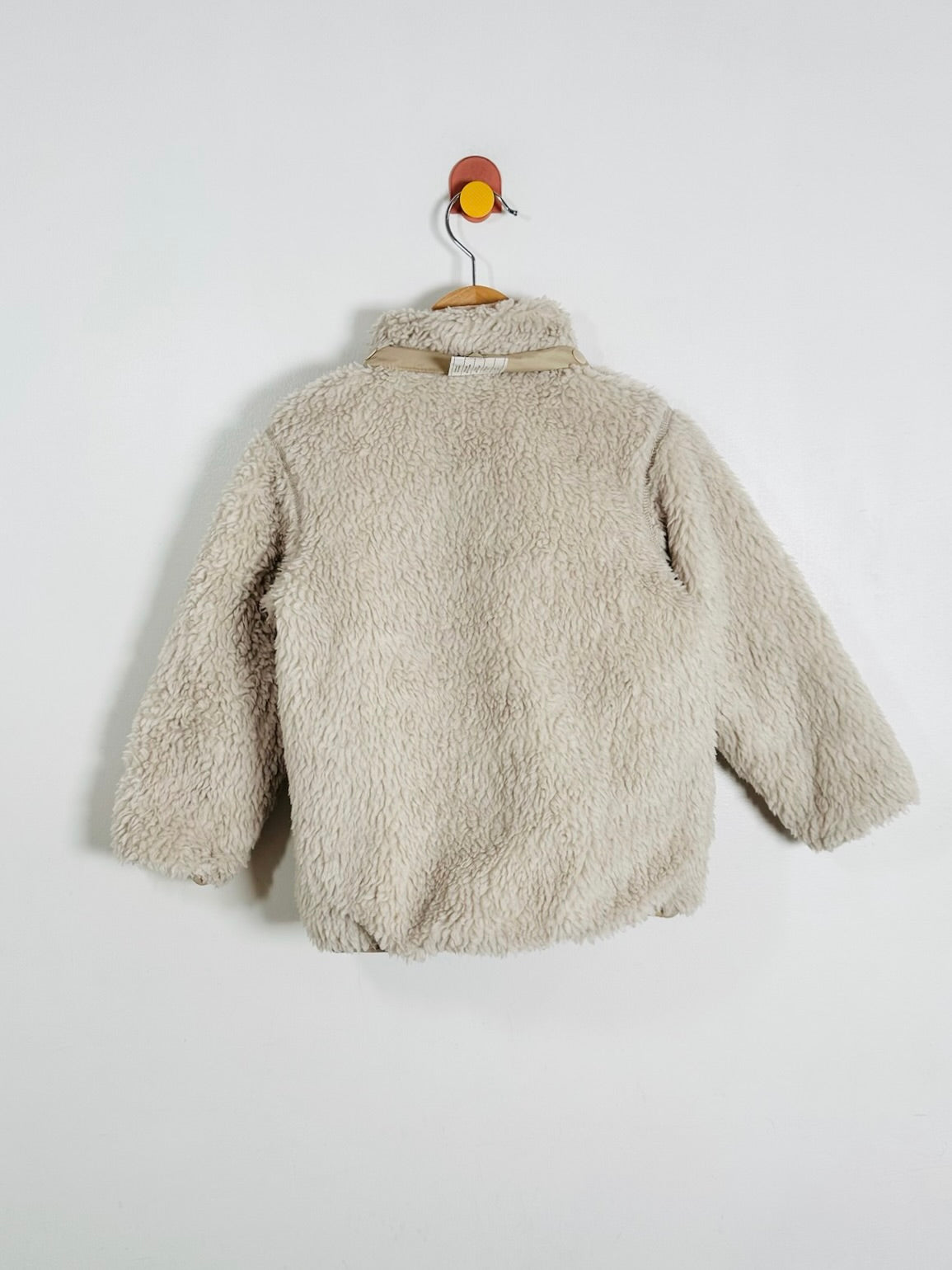 A beige sherpa fleece jacket hanging on a wooden hanger with a decorative orange and yellow hook against a plain white wall.