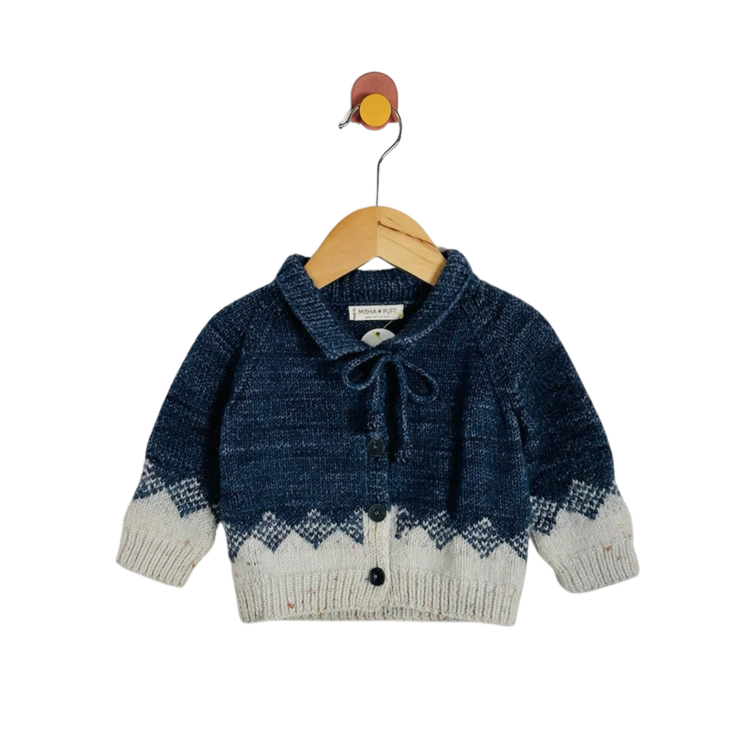 A baby knit cardigan sweater in navy blue and cream, featuring a tie-neck bow and button closure, hanging on a wooden hanger against a white background.