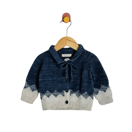 A baby knit cardigan sweater in navy blue and cream, featuring a tie-neck bow and button closure, hanging on a wooden hanger against a white background.