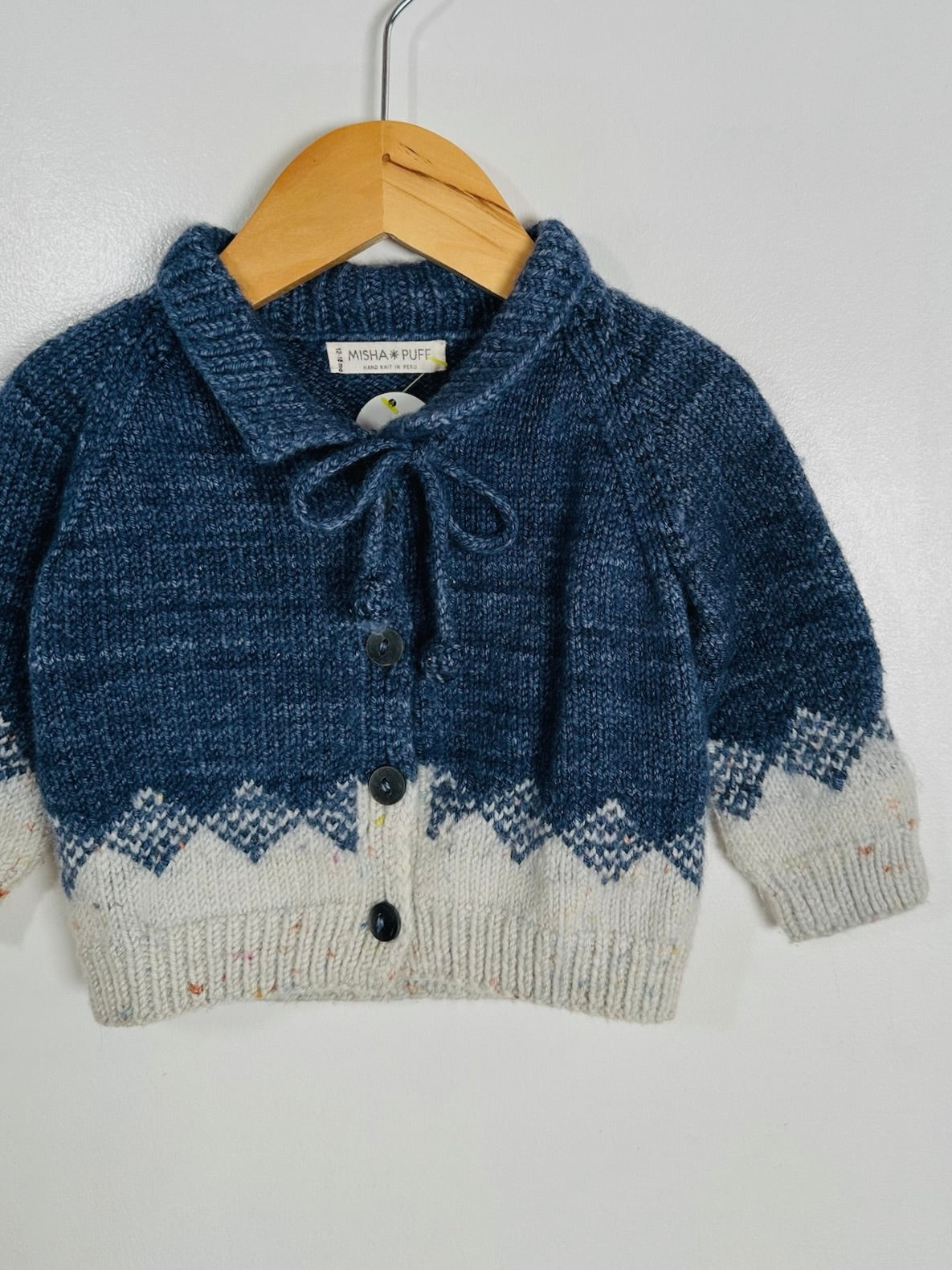 A baby knit cardigan sweater in navy blue and cream, featuring a tie-neck detail, button closure, and a Fair Isle patterned hem, hanging on a wooden hanger against a plain light gray wall.