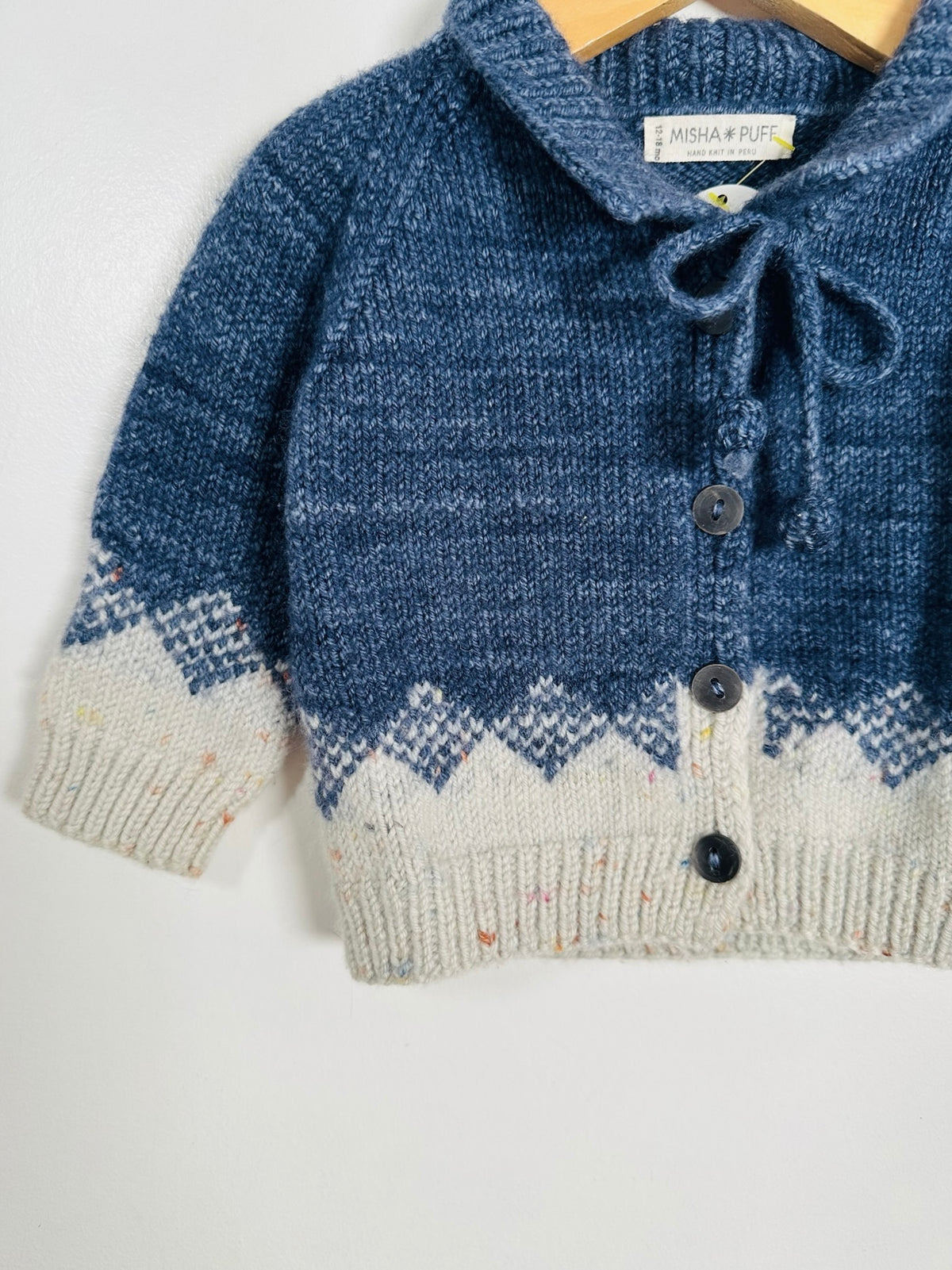 A close-up of a knitted baby cardigan sweater in blue and cream with a Fair Isle pattern, hanging on a wooden hanger against a plain white wall.