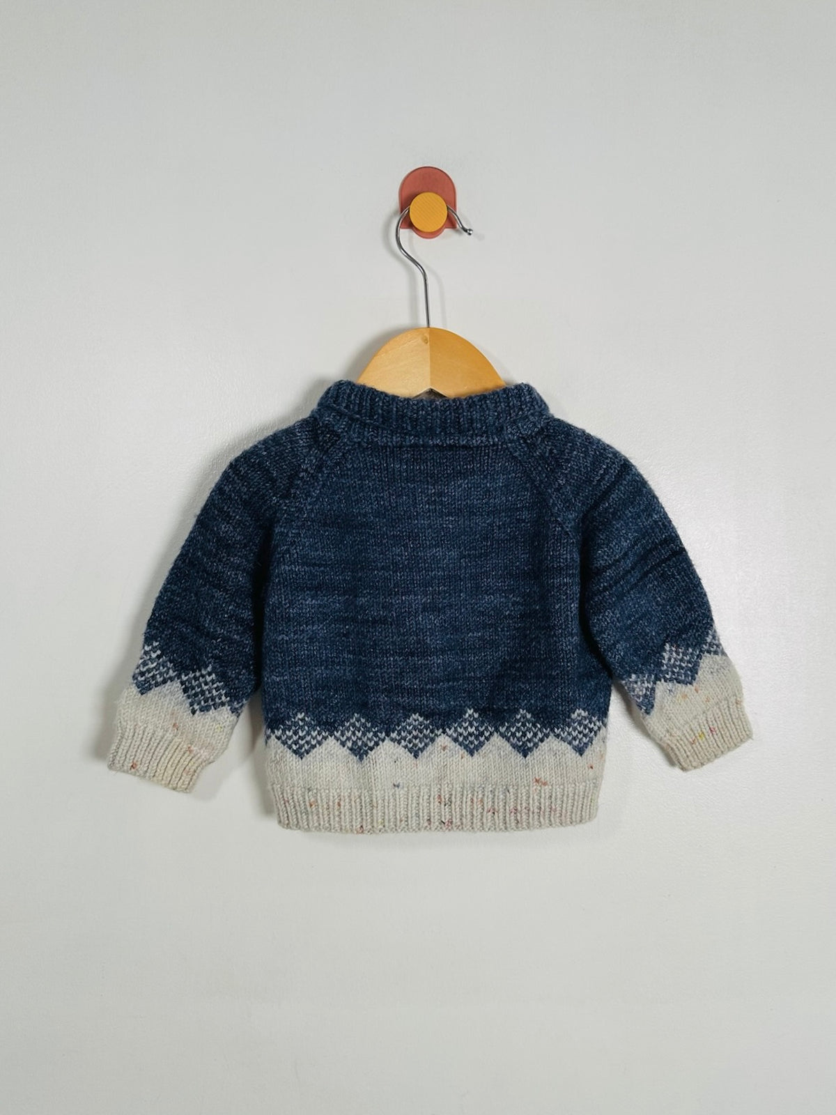 A baby knit sweater shown from the back, hanging on a wooden hanger against a plain light gray wall. The sweater is dark blue with a cream-colored geometric pattern along the hem and cuffs.