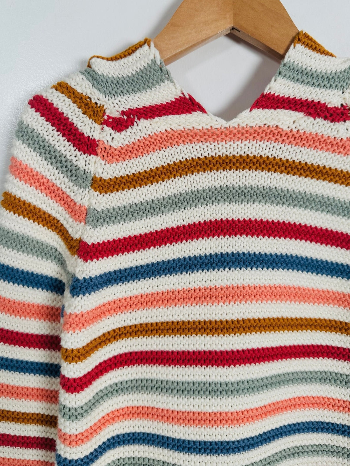 A close-up of a hand-knit sweater with horizontal stripes in red, blue, orange, brown, and cream, hanging on a wooden hanger against a light gray wall.