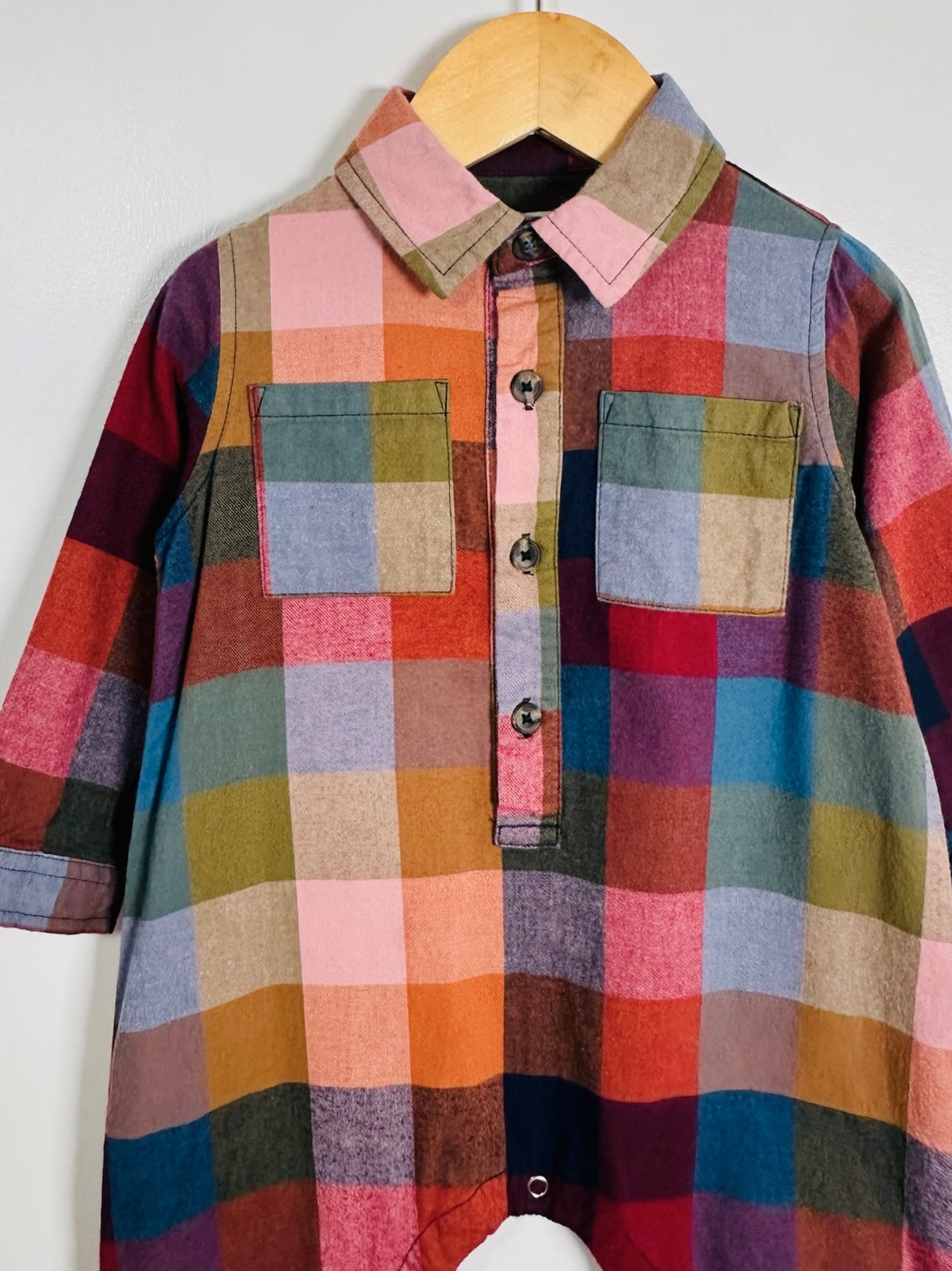A multicolor plaid button-up shirt with a pointed collar and two chest pockets, hanging on a wooden hanger against a plain light gray wall.