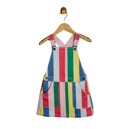 A children's sleeveless pinafore-style dress with colorful vertical stripes in pink, blue, green, yellow, and white, hanging on a wooden hanger against a plain white background.