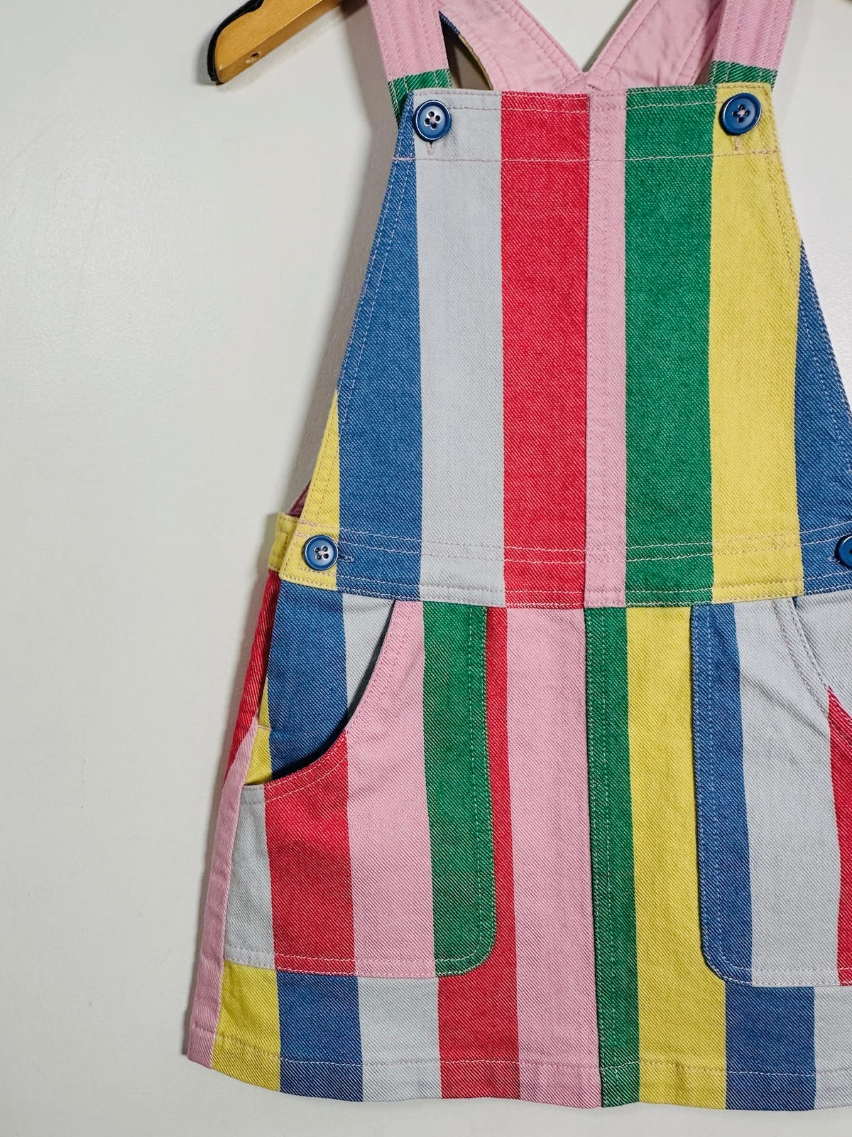 A children's striped overall dress with front pockets and blue buttons, hanging on a wooden hanger against a plain white wall.