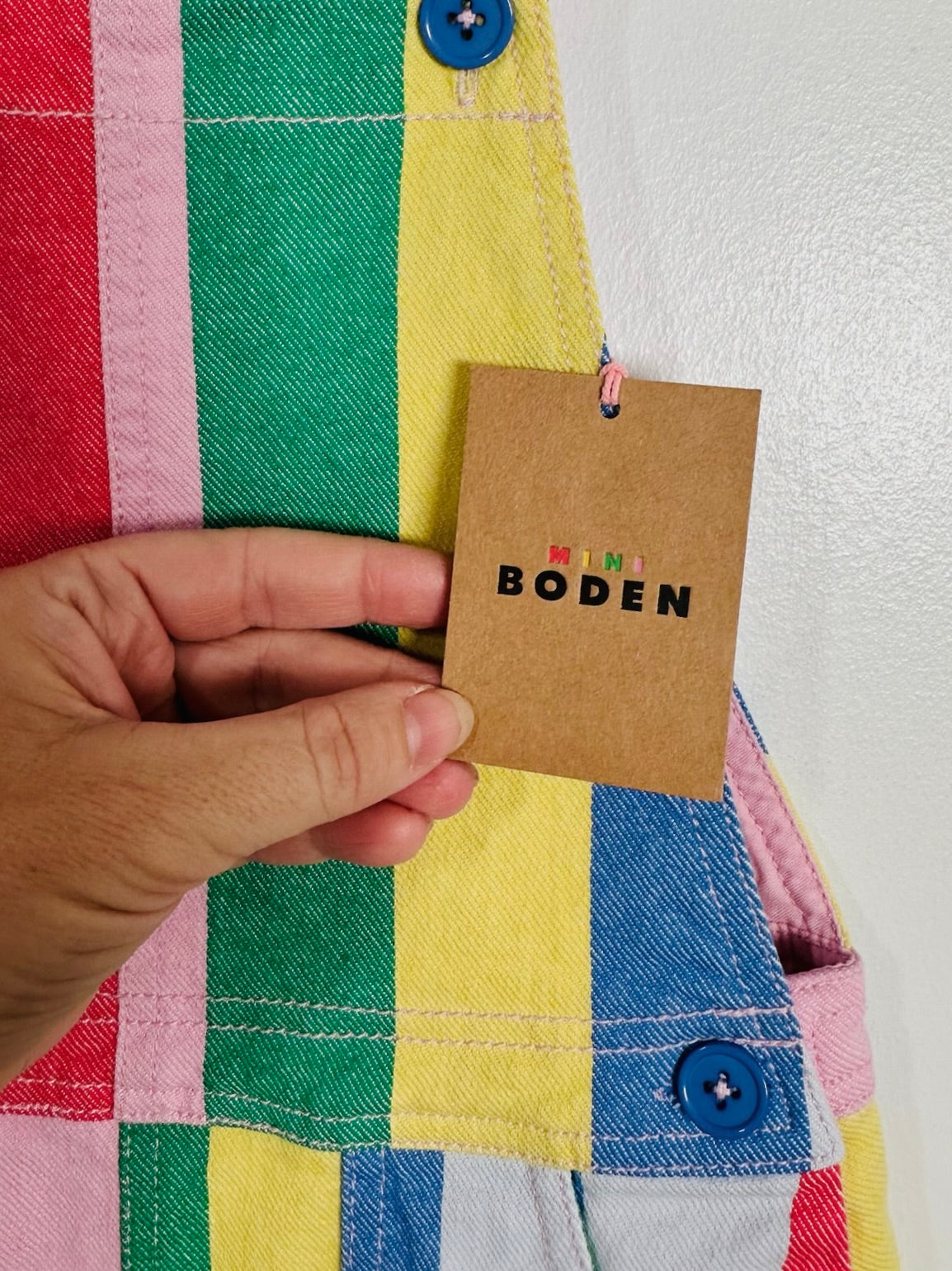 A close-up of a hand holding a brown paper tag that reads 'BODEN' in black letters, attached to a colorful striped denim garment with pink, green, yellow, and blue panels.