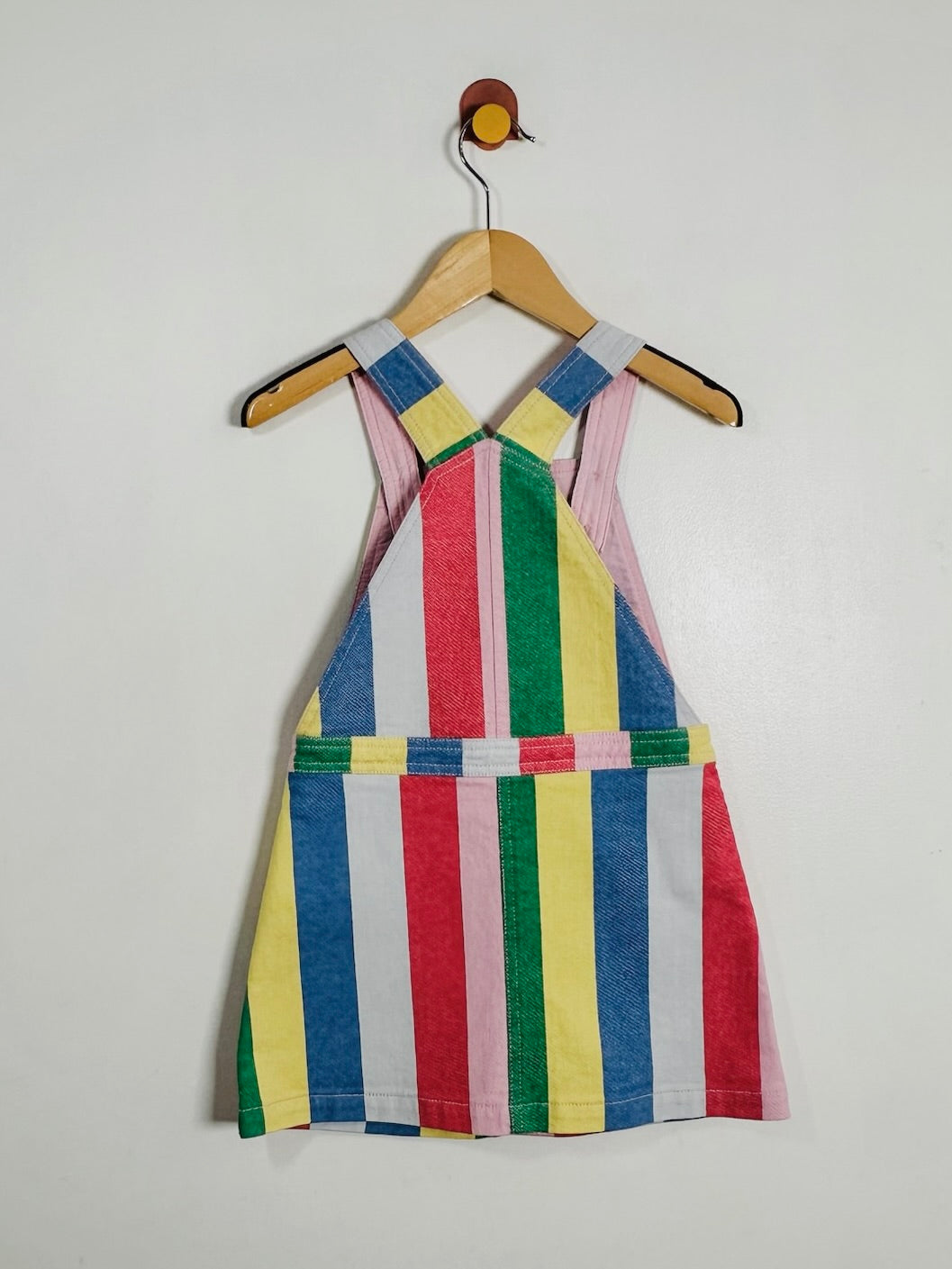 A children's sleeveless pinafore-style dress with vertical multicolor stripes in blue, red, green, yellow, pink, and white, hanging on a wooden hanger against a plain light wall.