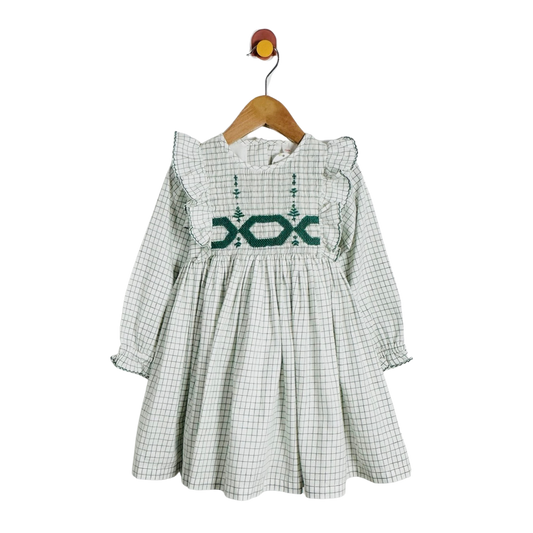 A long-sleeve baby dress with a green and white gingham pattern, featuring a dark green embroidered geometric design across the chest and ruffled shoulders, hanging on a wooden hanger against a white background.