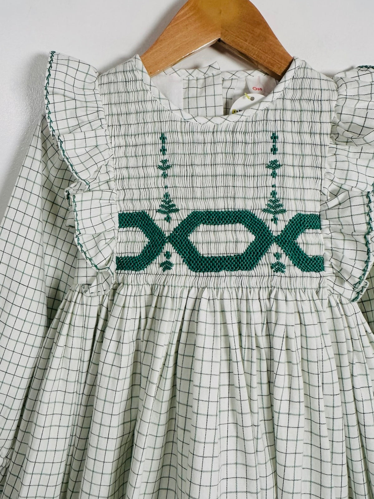 A children's long-sleeve dress with a white and green grid pattern, featuring dark green smocked geometric details across the chest and ruffled sleeves, hanging on a wooden hanger against a white wall.