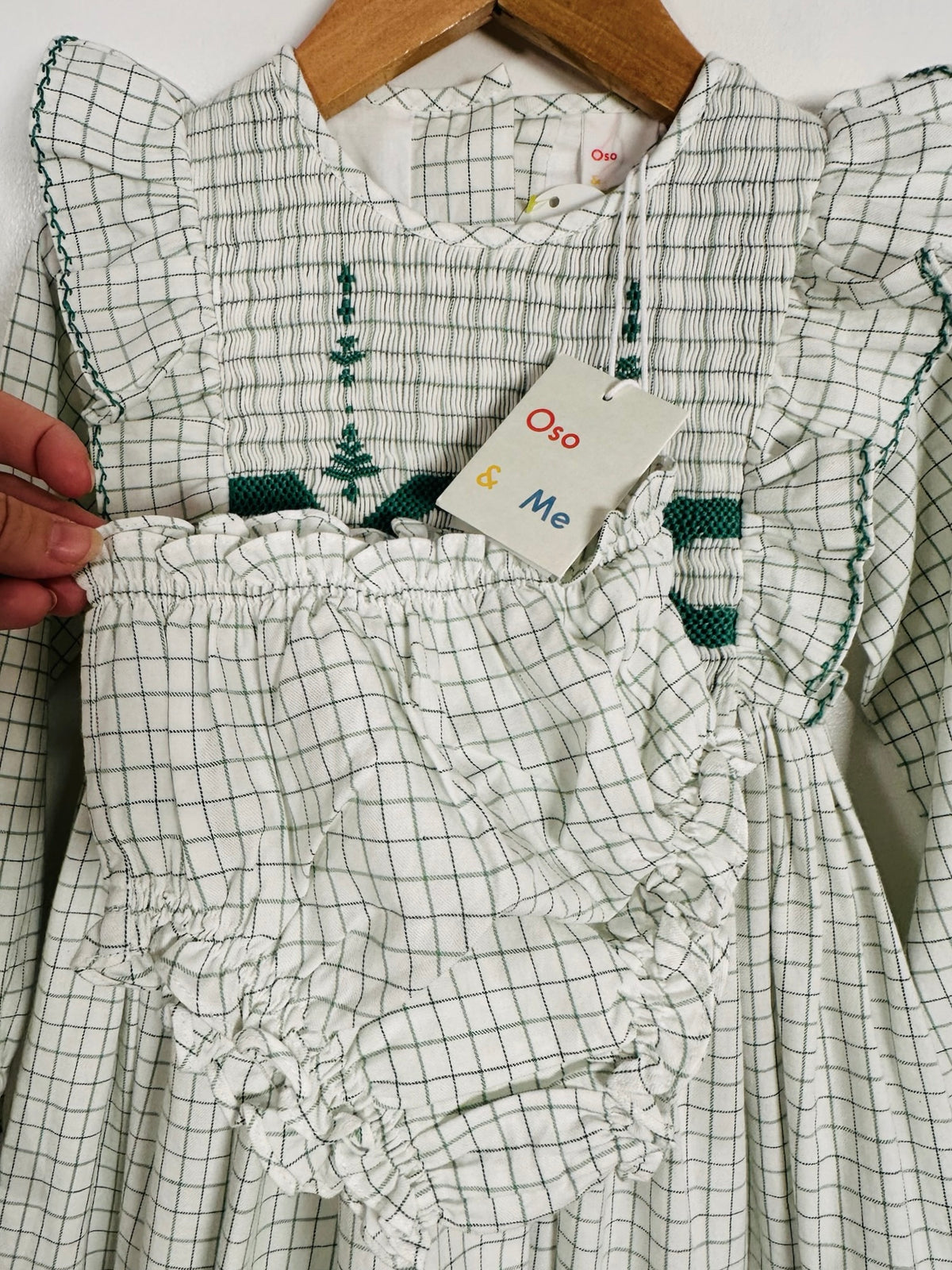 A children's smocked dress with ruffles, featuring a white and green gingham pattern, hanging on a wooden hanger. A hand is holding the ruffled hem to show its flexibility.
