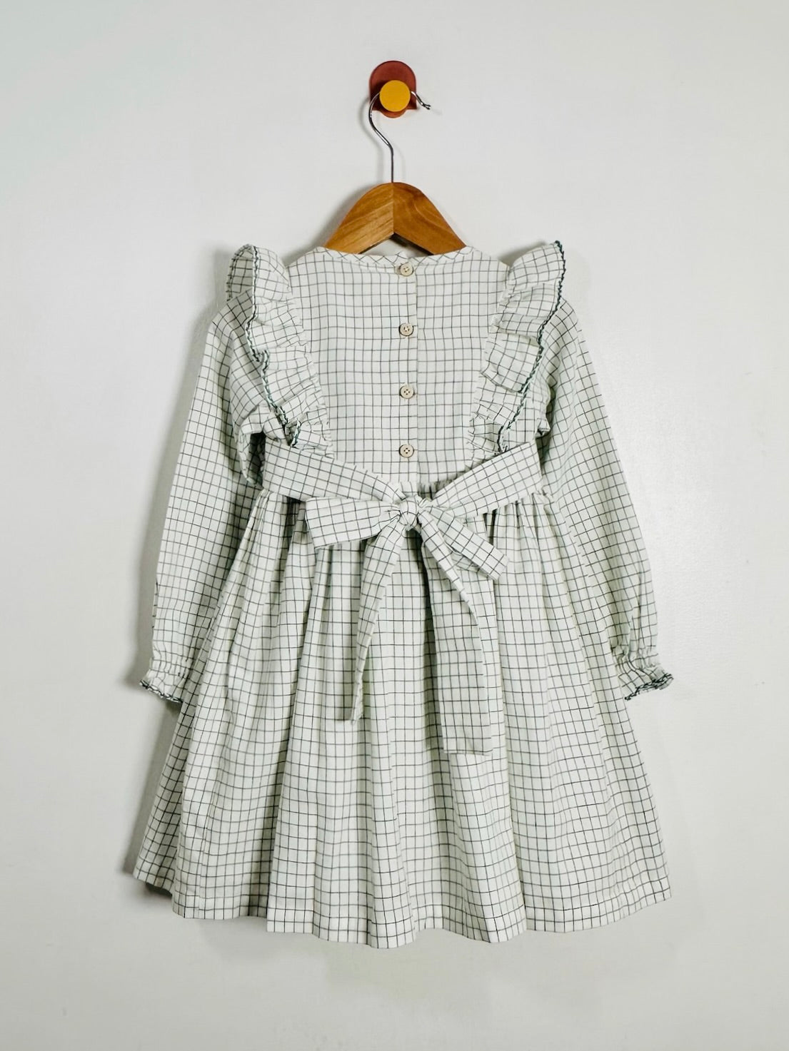 A long-sleeve children's dress with a green and white windowpane plaid pattern, featuring ruffled shoulders, a buttoned back, and a matching fabric tie belt at the waist, hanging on a wooden hanger against a plain light gray wall.