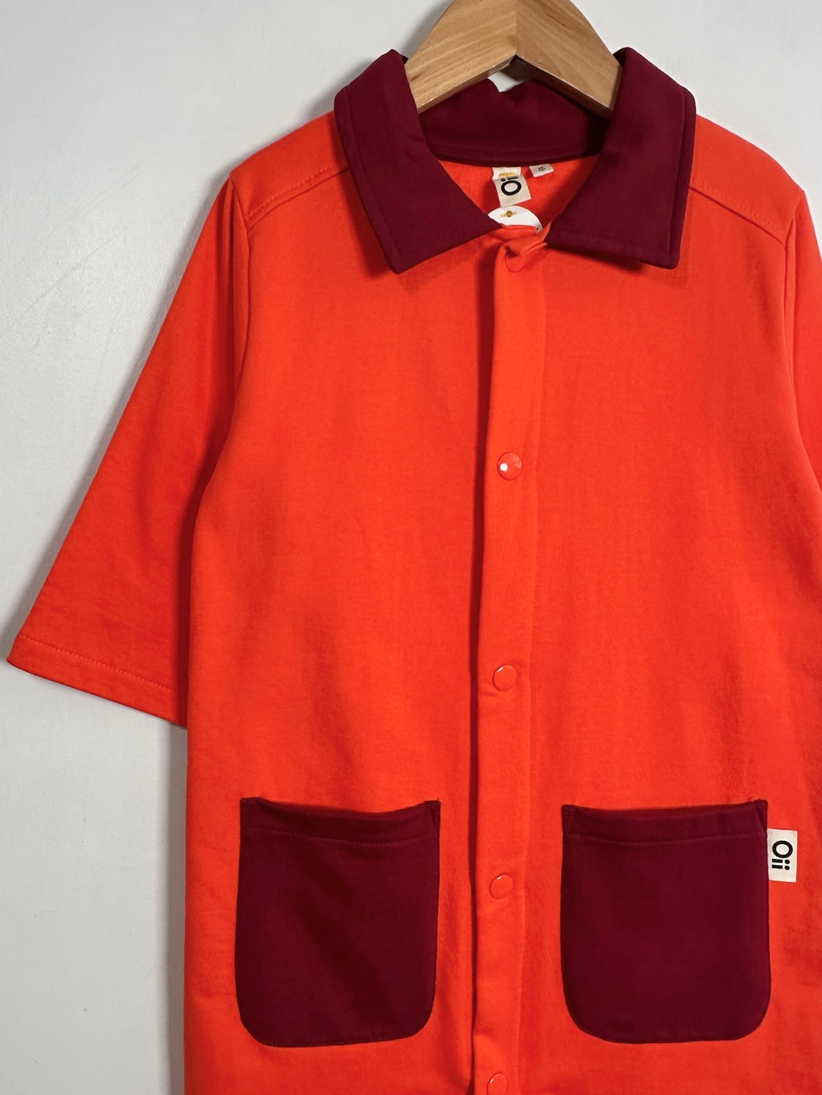 Bright orange short-sleeve button-front shirt jacket with contrasting dark red collar and patch pockets, hanging on a wooden hanger against a light gray wall.