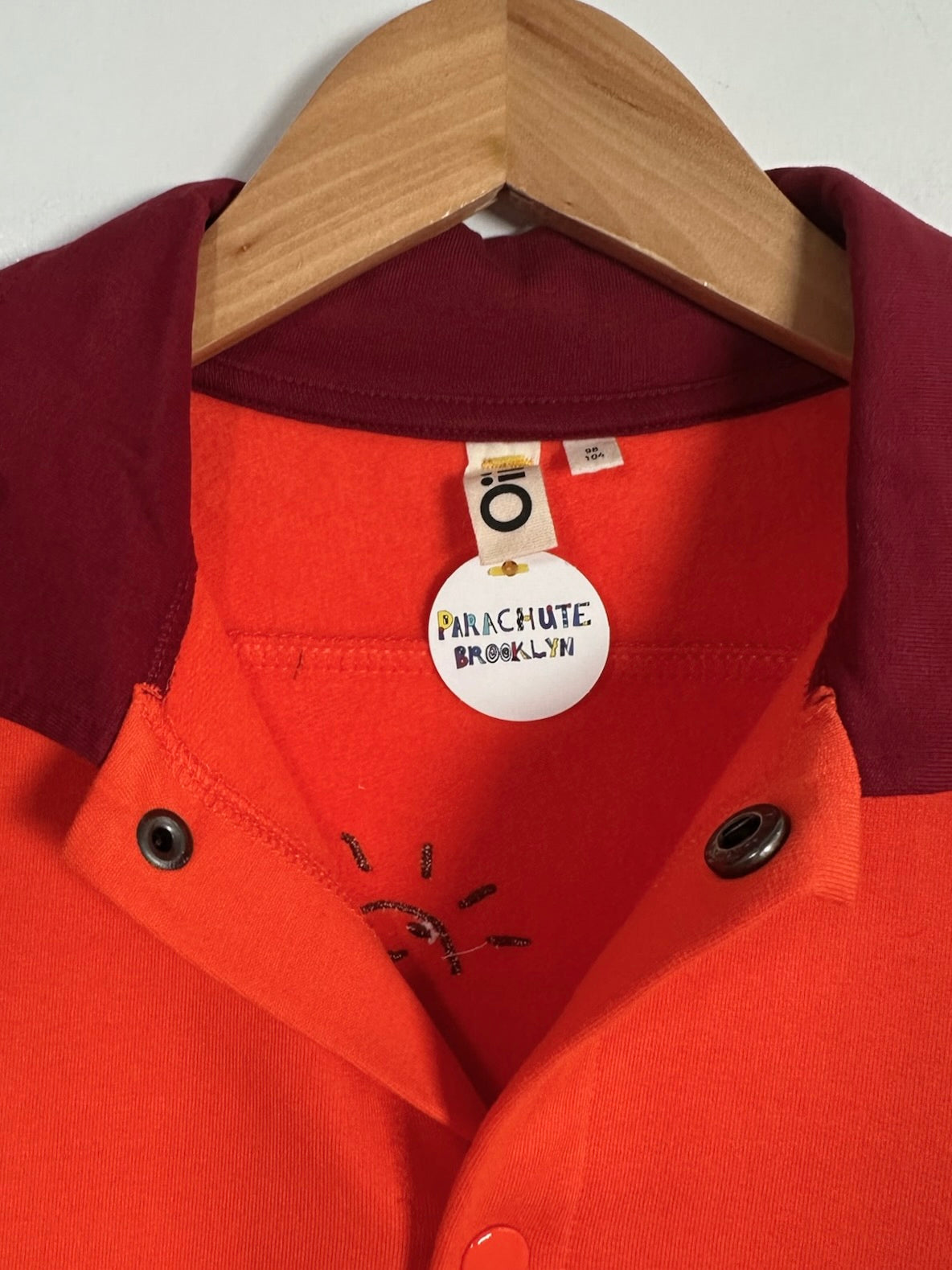 A close-up of a red-orange collared shirt on a wooden hanger, featuring a dark burgundy collar, a white circular neck label reading 'PARACHUTE BROOKLYN', and dark metal snap buttons.
