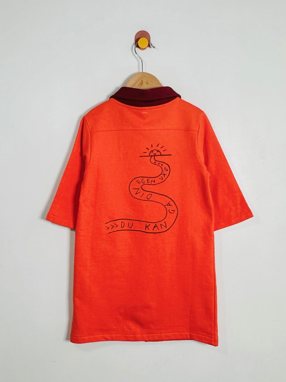 An orange short-sleeve shirt with a dark red collar, hanging on a wooden hanger against a light gray wall. The back of the shirt features a black hand-drawn snake graphic with the text 'DU KAN' and '>>>' written on it.