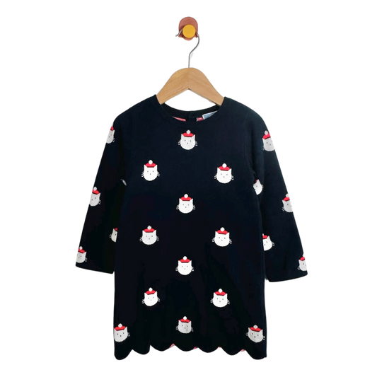 A blue long-sleeve dress with a scalloped hem, featuring a repeating pattern of white cartoon cat faces wearing red hats, hanging on a wooden hanger against a plain white background.