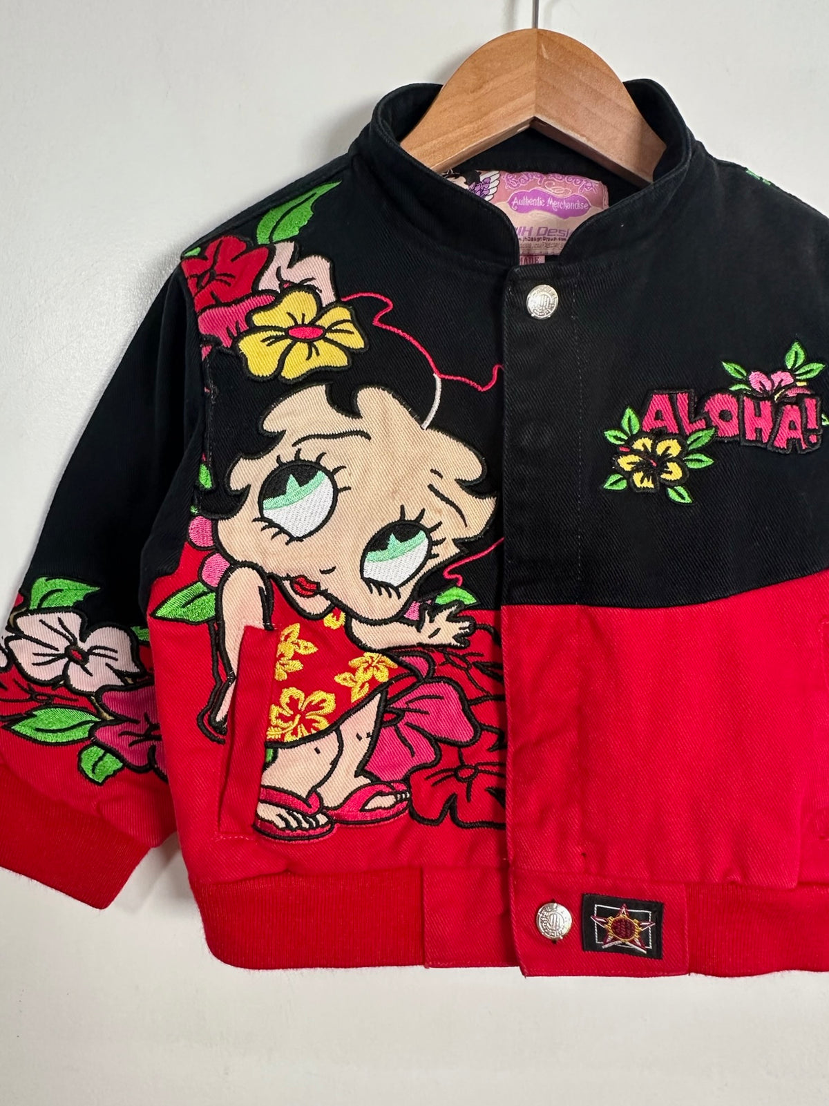 A vintage children's bomber jacket hanging on a wooden hanger against a plain white wall. The jacket features a black upper section and a red lower section, decorated with a large cartoon Betty Boop character on the left side, colorful floral embroidery, and 'ALOHA!' text on the right side.
