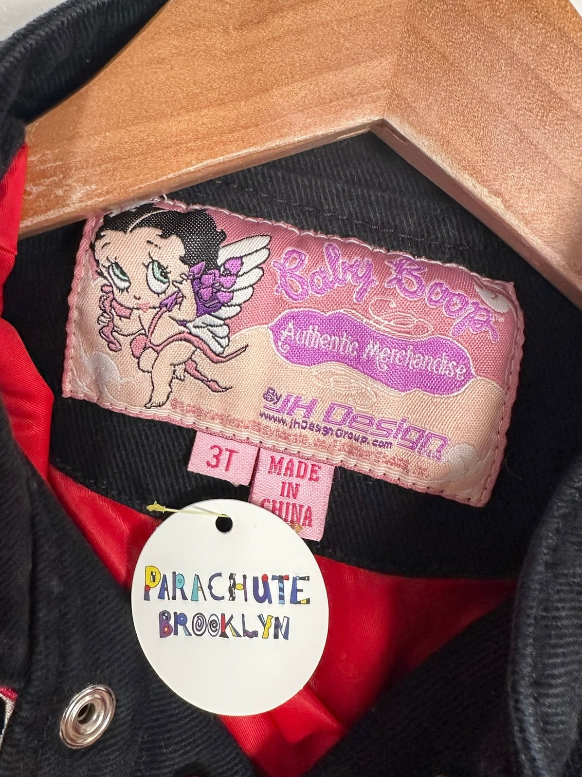 Close-up of a vintage black jacket with a red lining, featuring a pink 'Baby Boop' authentic merchandise label and a white 'Parachute Brooklyn' hang tag.