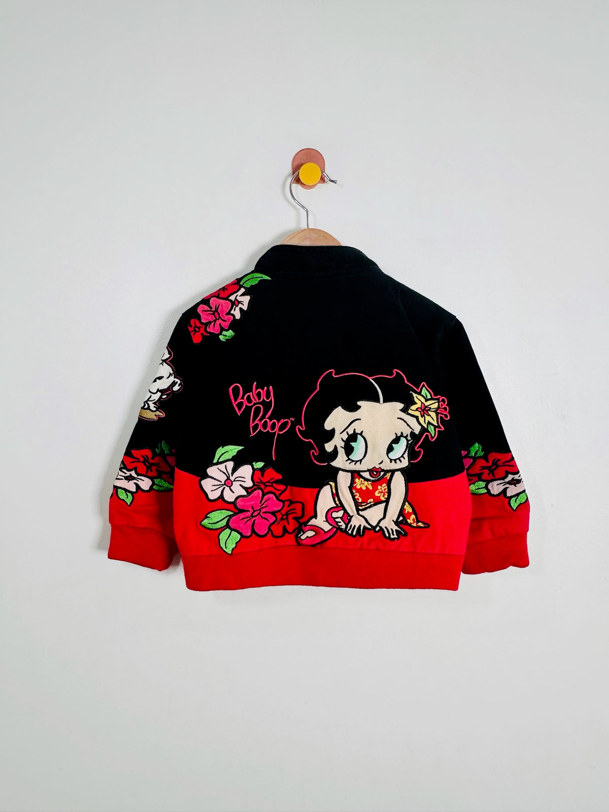 A vintage black and red bomber jacket featuring a cartoon Betty Boop design on the back, hanging on a wooden hanger against a plain light gray wall.