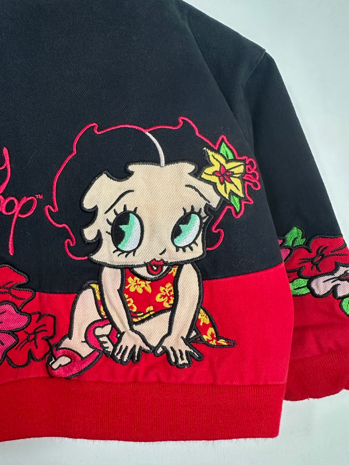 A close-up of a black and red jacket featuring a large embroidered cartoon Betty Boop character on the front, with floral embroidery on the sleeve.