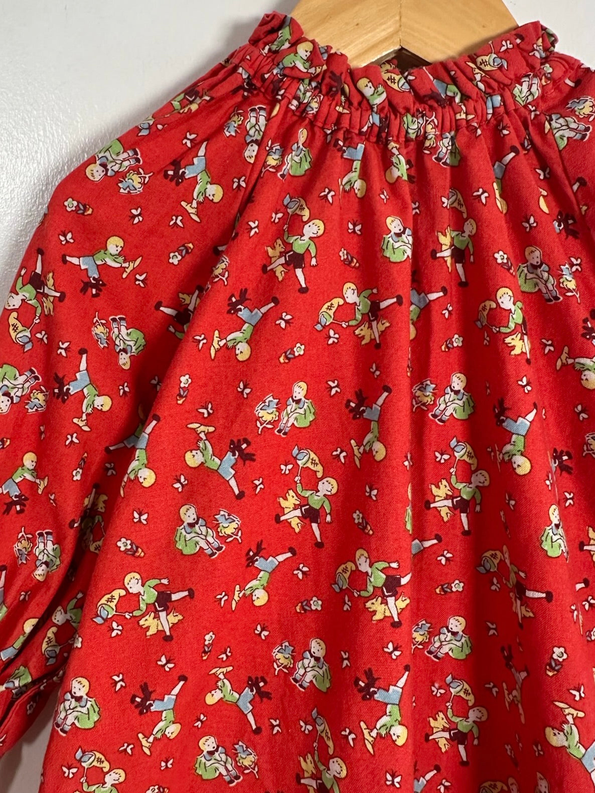A red fabric garment, likely a dress or long-sleeve top, featuring an all-over print of cartoon figures, hanging on a wooden hanger against a plain white wall.