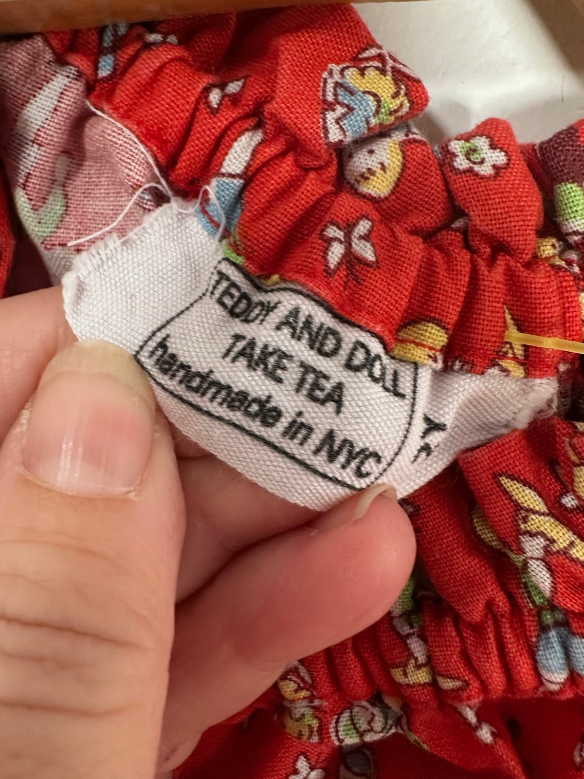 Close-up of a red fabric garment with a white sewn-in label reading 'TEDDY AND DOLL TAKE TEA handmade in NYC', held by a person's fingers.