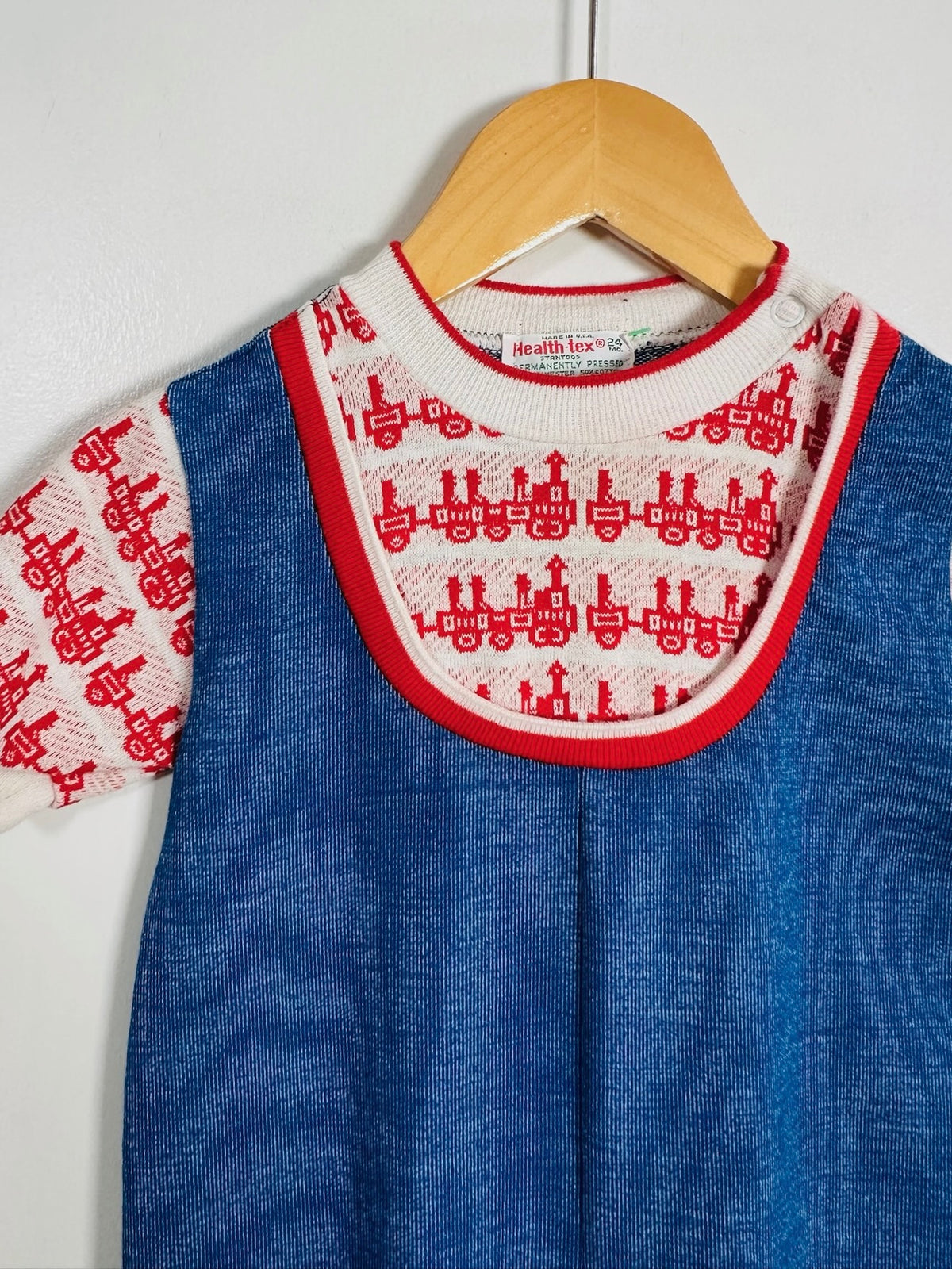 A blue knit baby dress with a red-trimmed white bib-style collar and short sleeves featuring a red train pattern, hanging on a wooden hanger against a white wall.