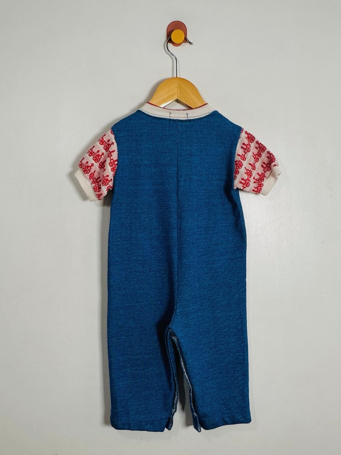 A baby romper hanging on a wooden hanger against a plain light gray wall. The romper is primarily blue with short sleeves featuring a red and white patterned print and a white collar.