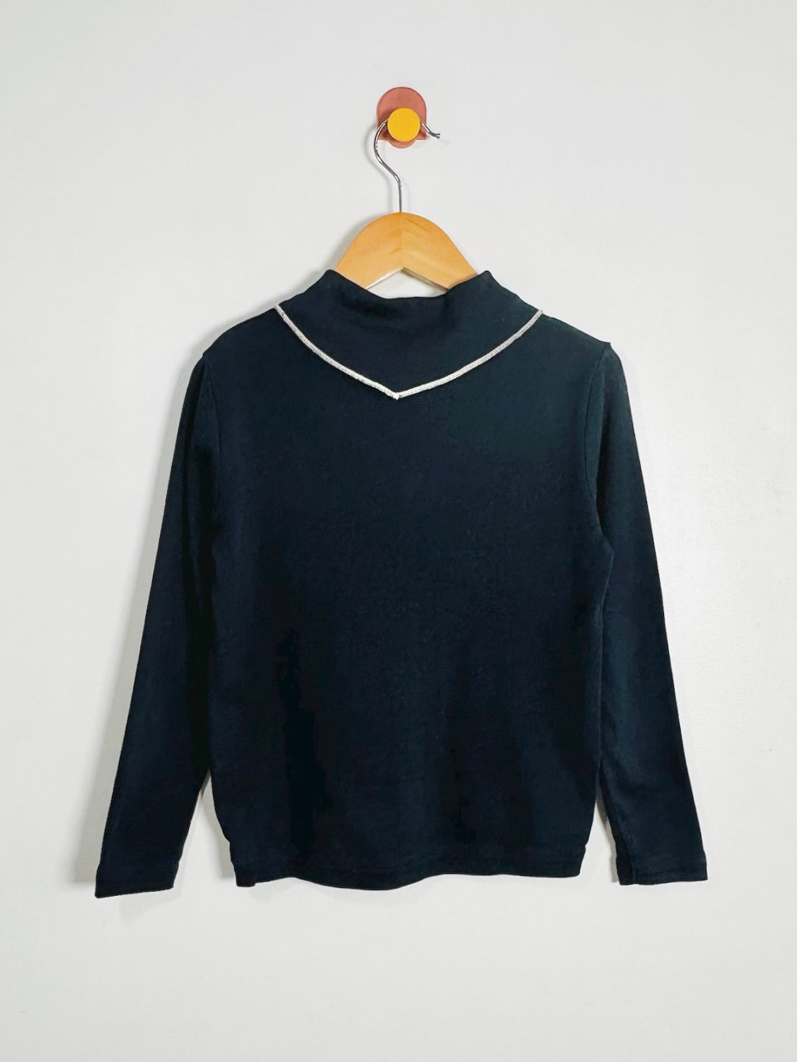 A dark navy blue long-sleeve mock neck sweater with a thin white contrast trim along the V-neckline, hanging on a wooden hanger against a plain light gray wall.