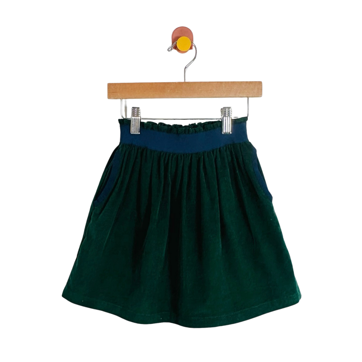 A dark green corduroy skirt with a navy blue elastic waistband, hanging from a wooden hanger with metal clips against a plain white background.