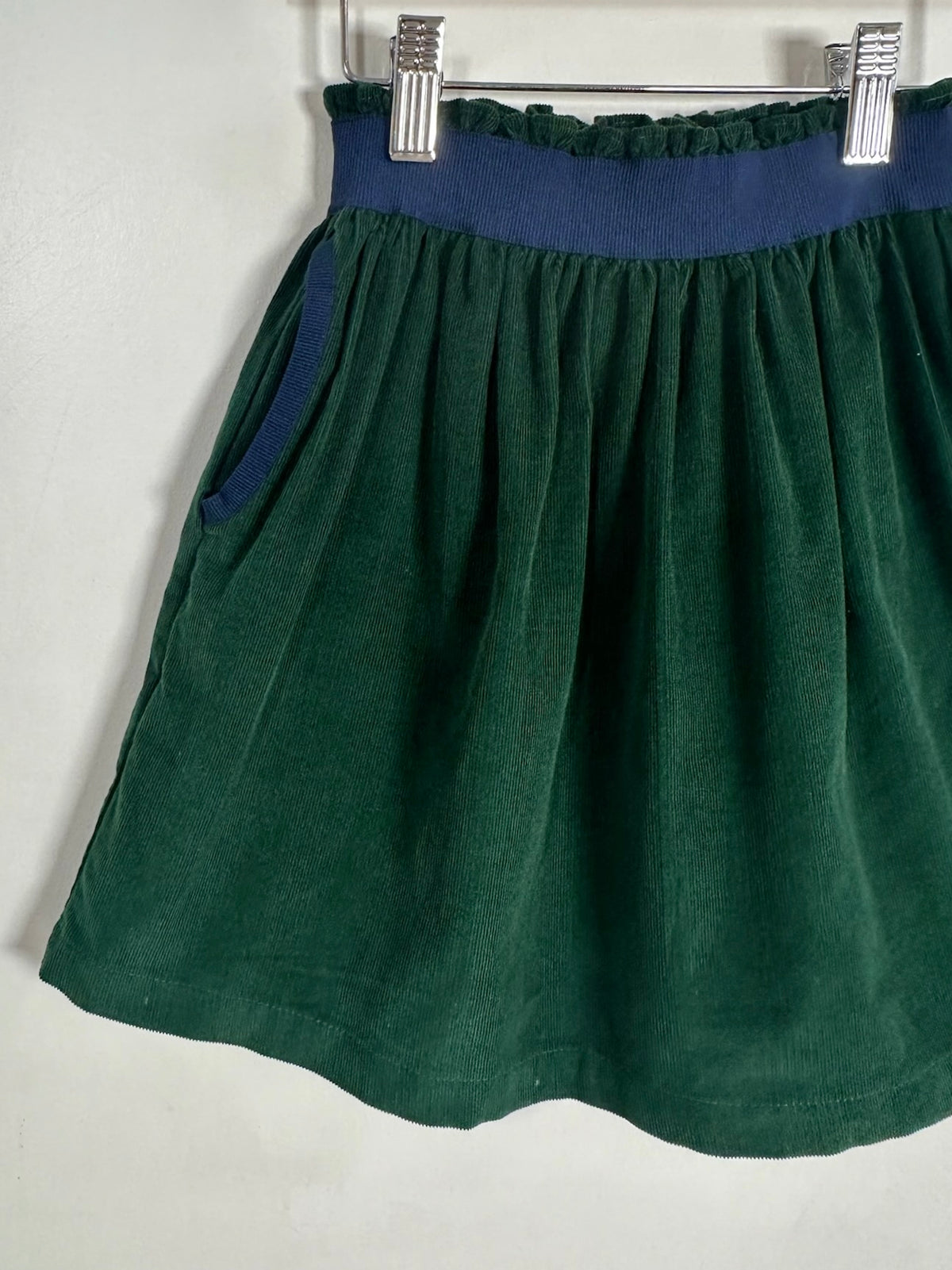 A dark green skirt with a contrasting navy blue waistband, hanging from a metal hanger against a plain white wall.