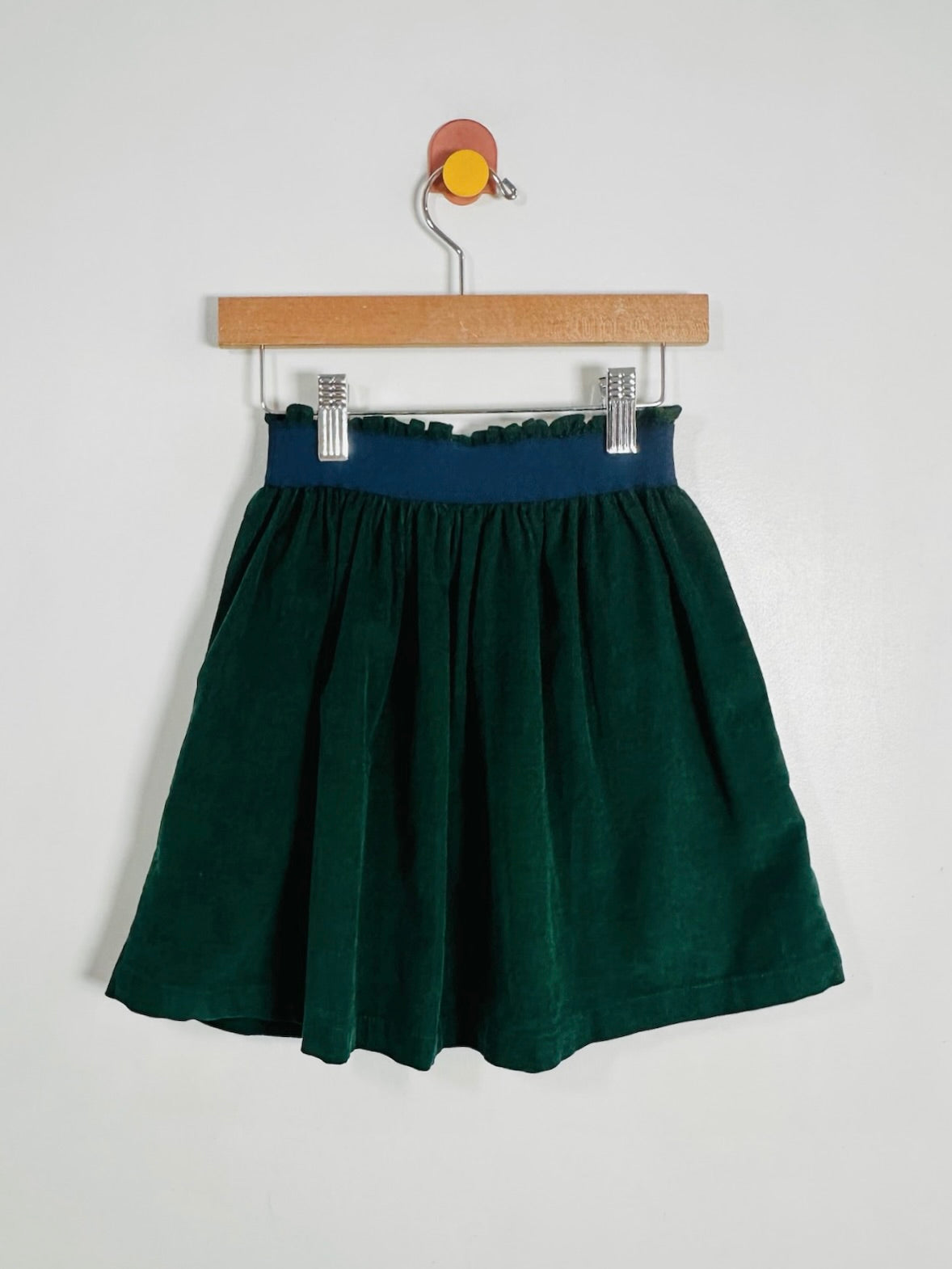 A dark green pleated skirt with a navy blue elastic waistband, hanging from a wooden hanger with silver clips against a plain white wall.