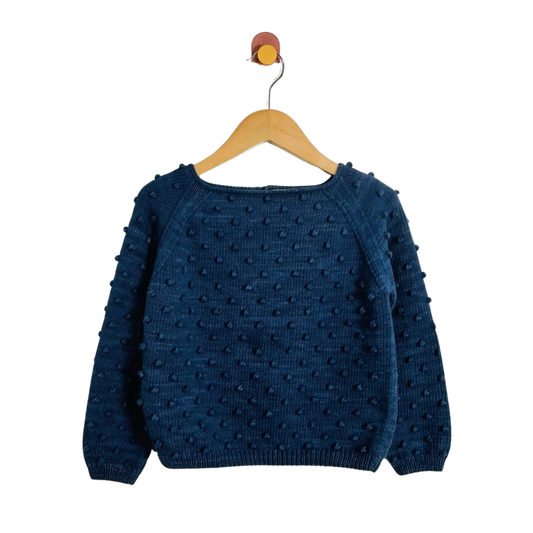 A dark blue textured knit sweater with raised pom-pom details, hanging on a wooden hanger against a plain white background.