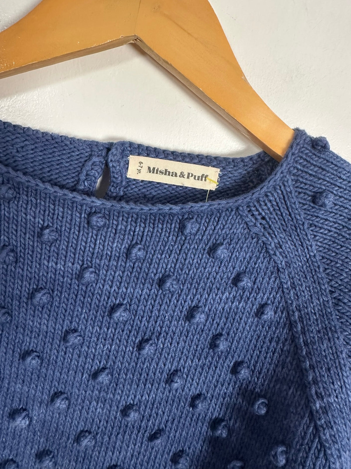 A close-up of a blue knit sweater with a textured bobble pattern, hanging on a wooden hanger against a white wall. The brand label 'Misha & Puff' is visible at the neckline.
