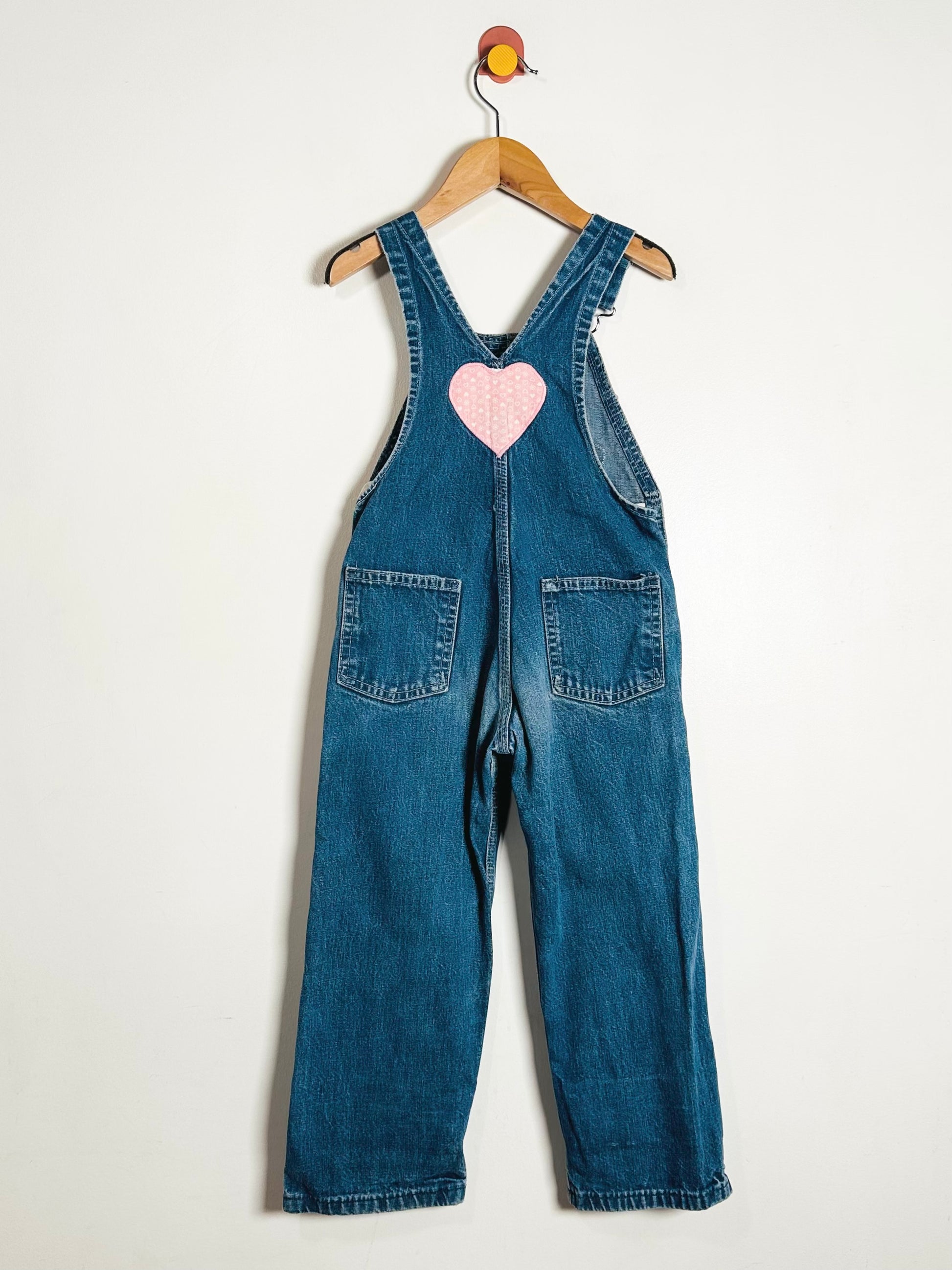 Blue denim overalls with a pink heart on a hanger against a white background
