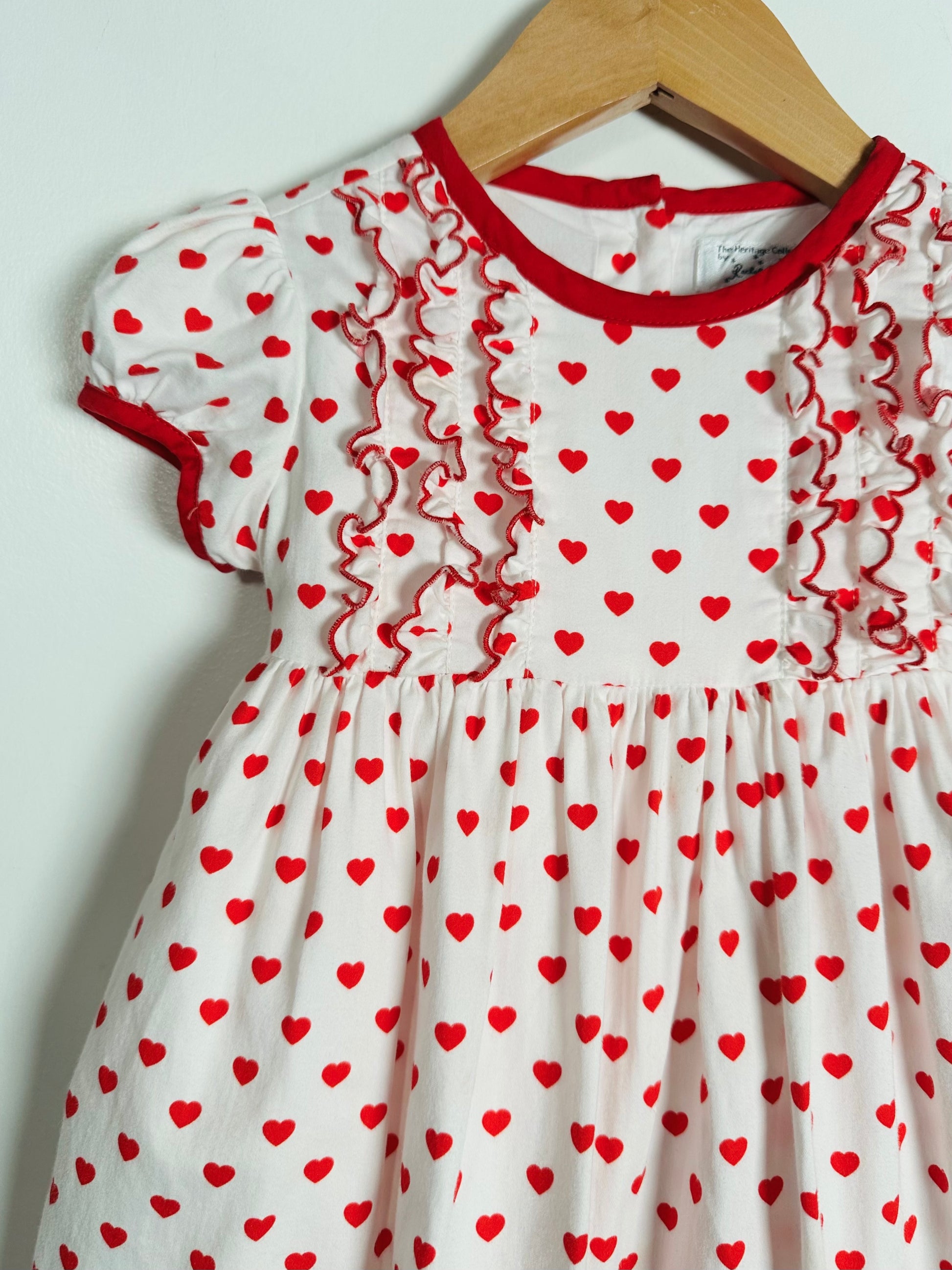 White dress with red heart pattern and ruffled trim on a hanger against a light gray background