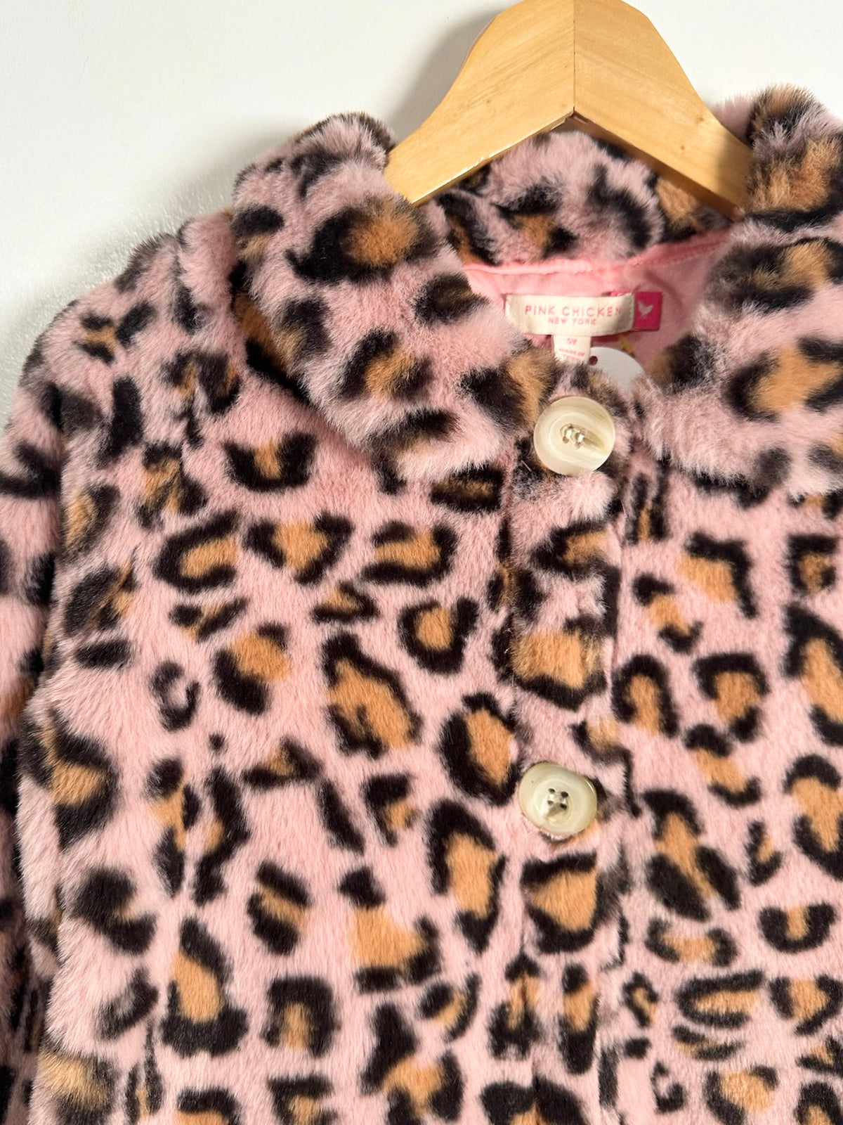 Pink faux fur jacket with a leopard print pattern, featuring a high collar and front buttons, hanging on a wooden hanger against a plain white wall.