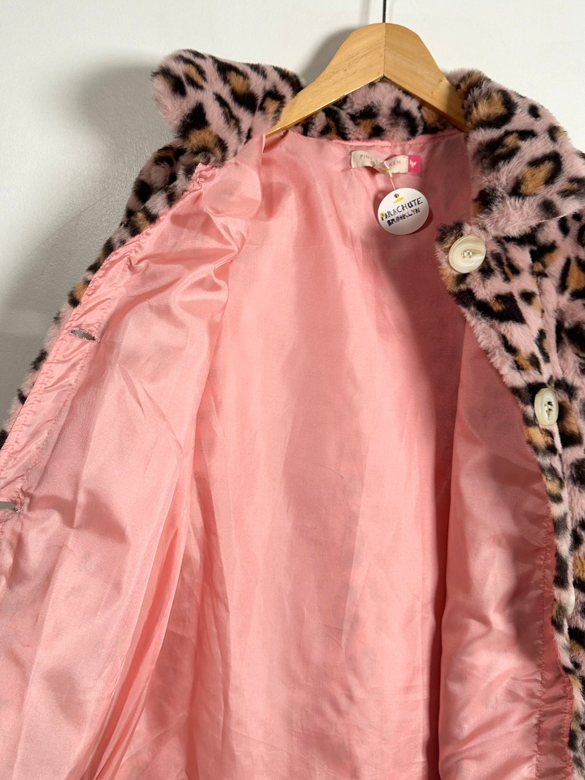 A pink satin-lined coat with a leopard-print faux fur collar, hanging on a wooden hanger against a plain white wall.