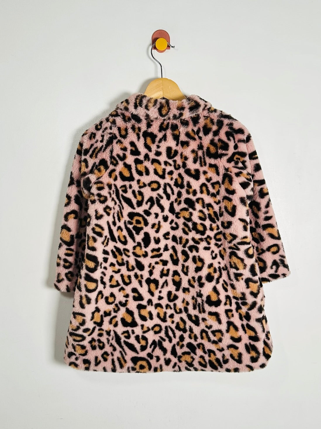 A pink faux fur jacket with a leopard print pattern, hanging on a wooden hanger against a plain white wall.